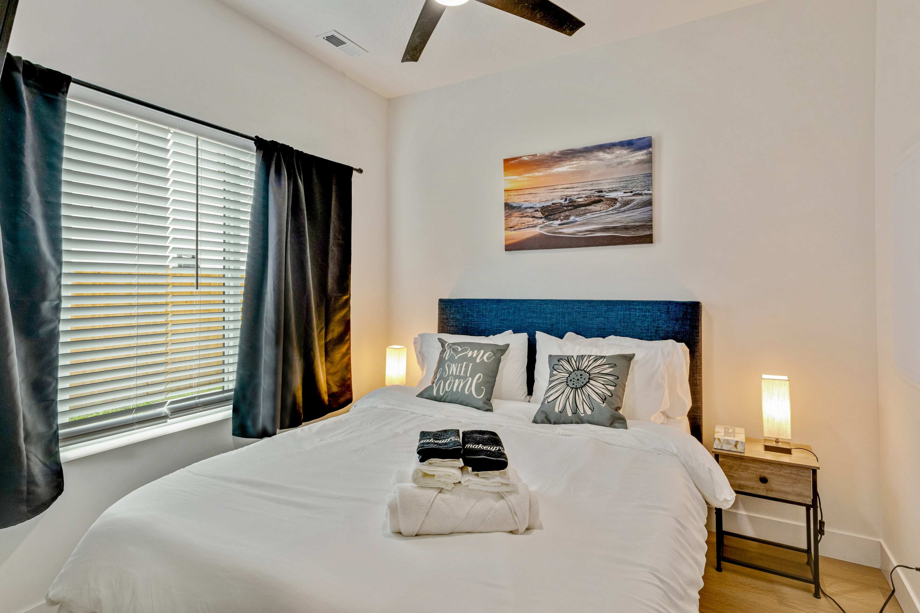 A neatly arranged bedroom features a white bed with decorative pillows and a beach-themed photograph above the headboard, alongside a nightstand with lamps and a stack of towels.