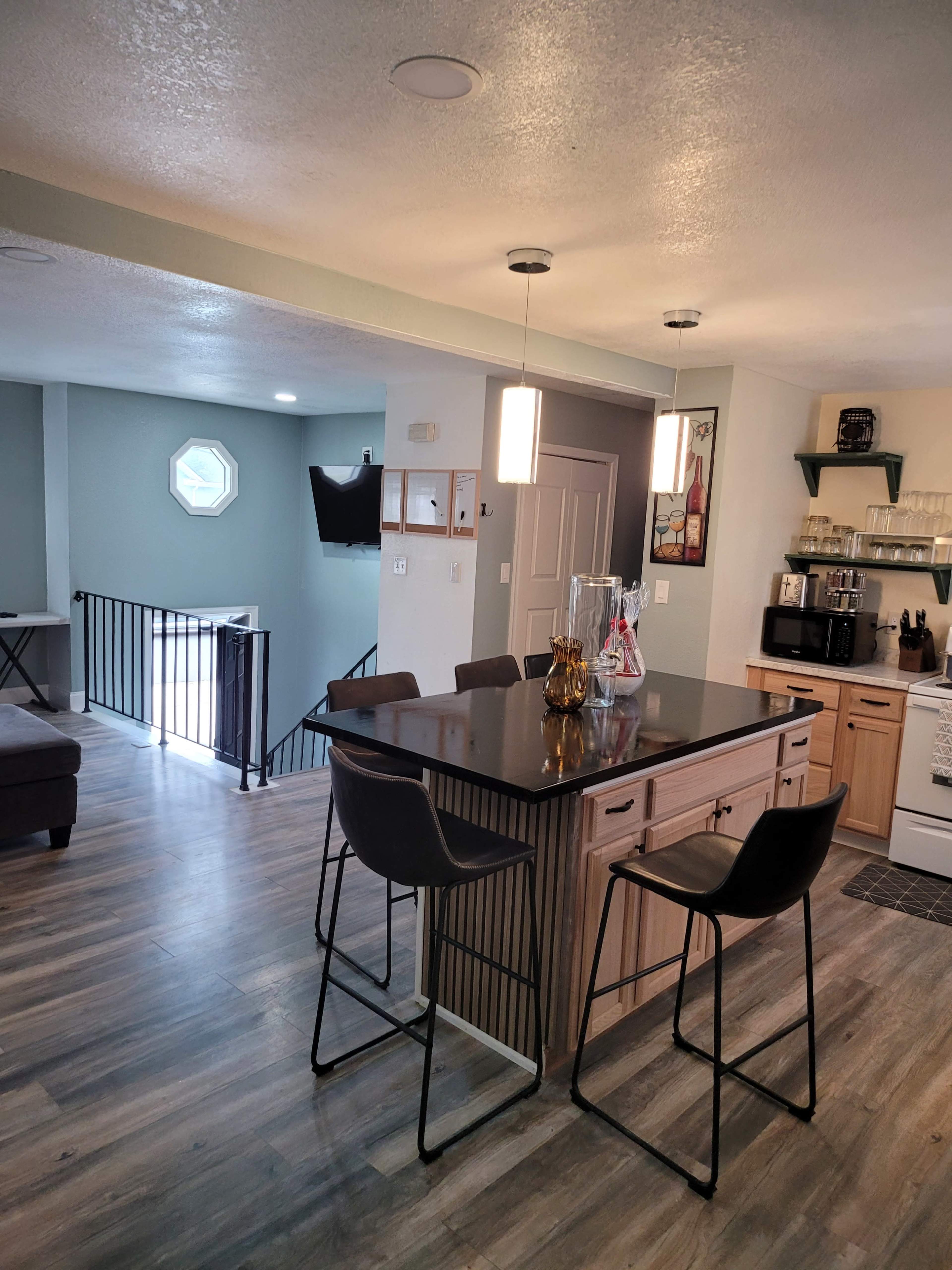 The image shows a modern kitchen with a central island, bar stools, and a living area visible in the background.