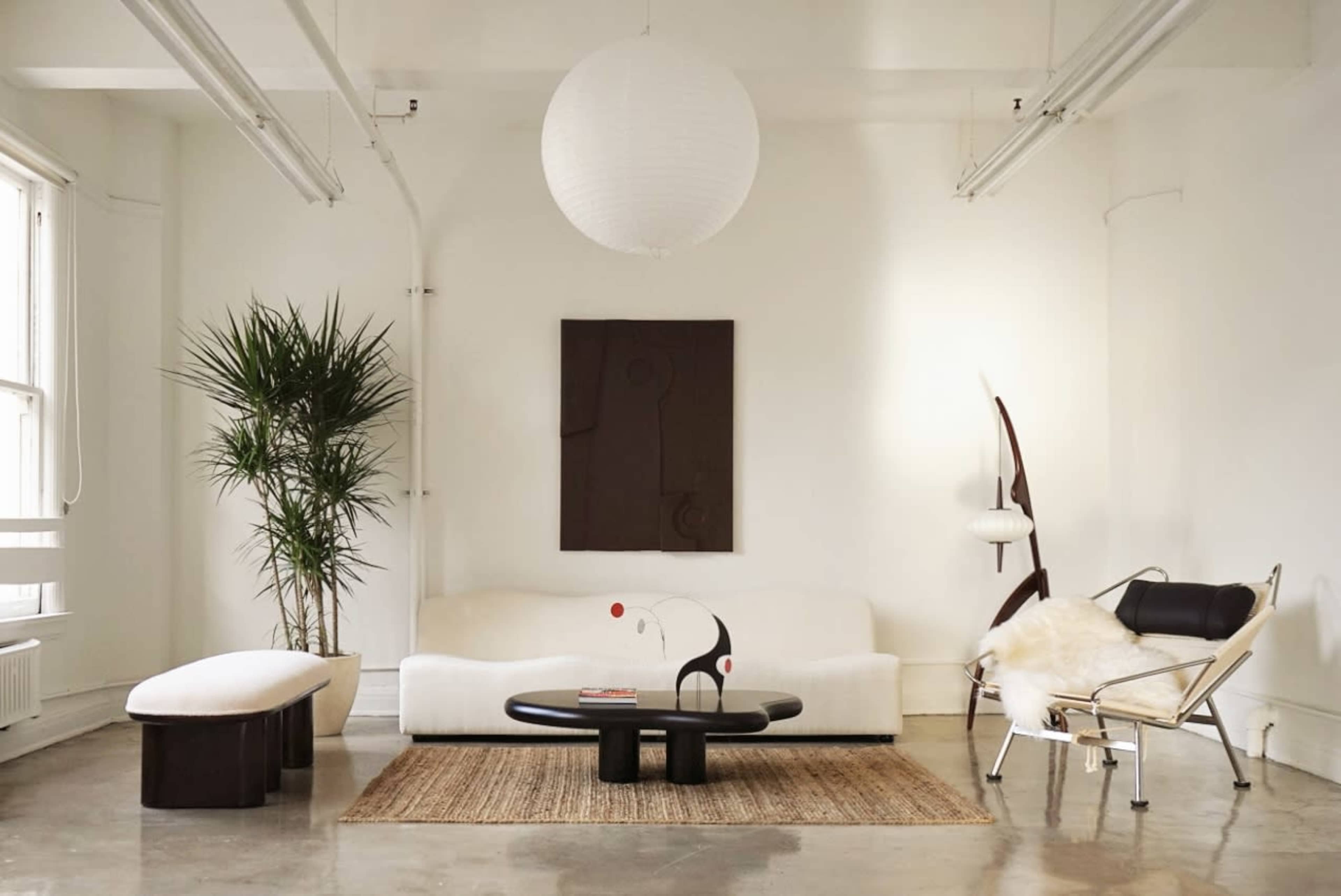 A bright living room features a white sofa, a round black coffee table, a textured rug, and a tall plant, complemented by modern art on the wall.