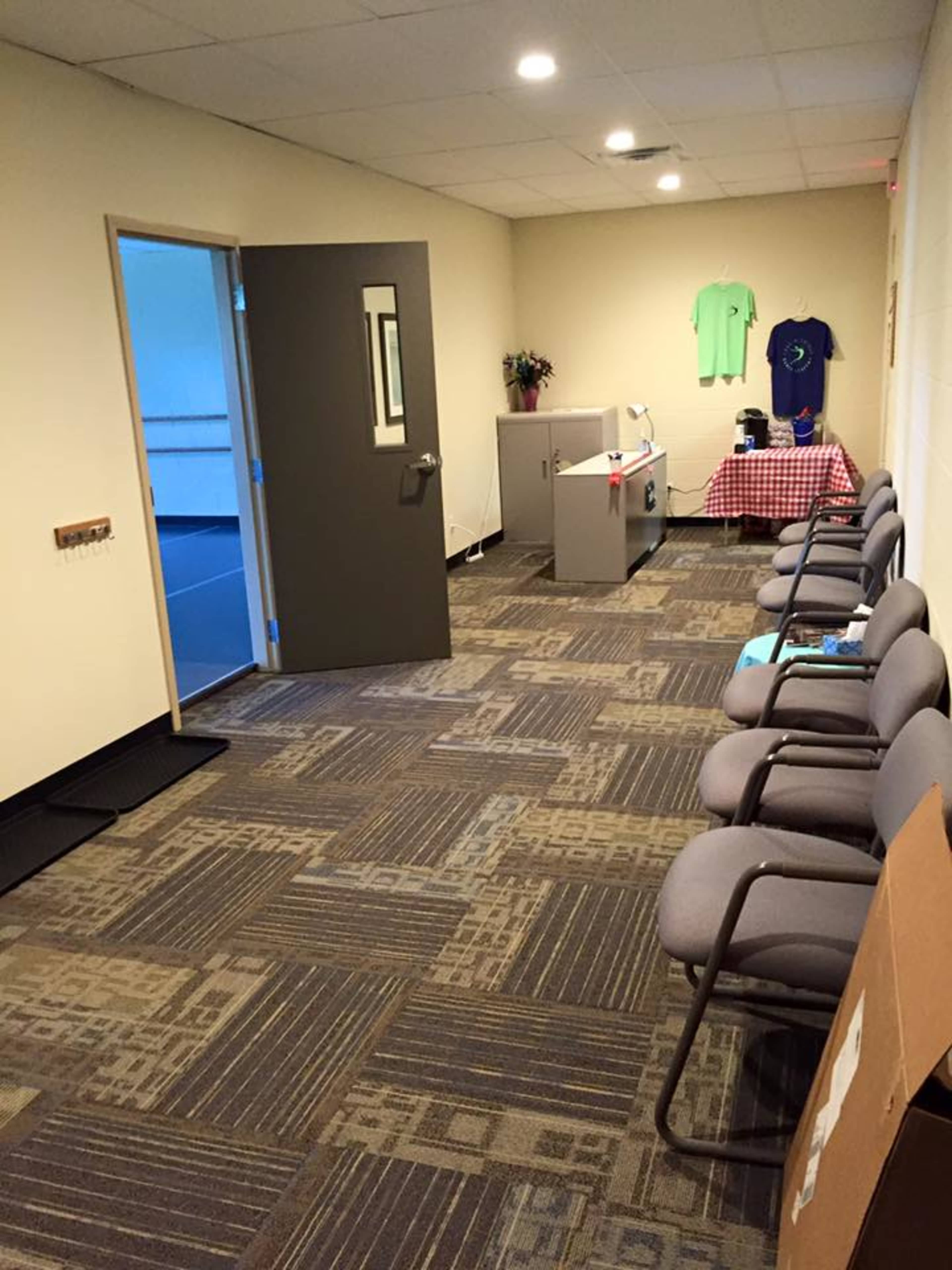 A hallway with gray chairs along one side, a desk with a checkered tablecloth at the end, and a door leading to another room.