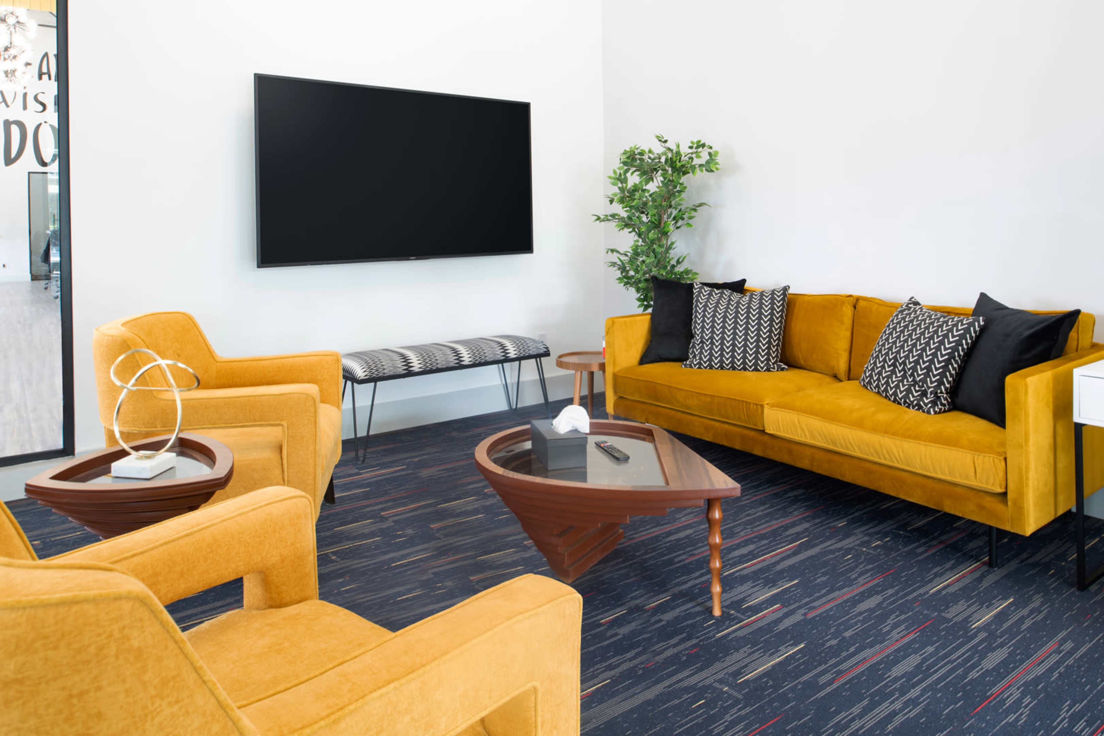 A brightly lit lounge area features a yellow sofa and armchairs, a glass coffee table, and a wall-mounted television.