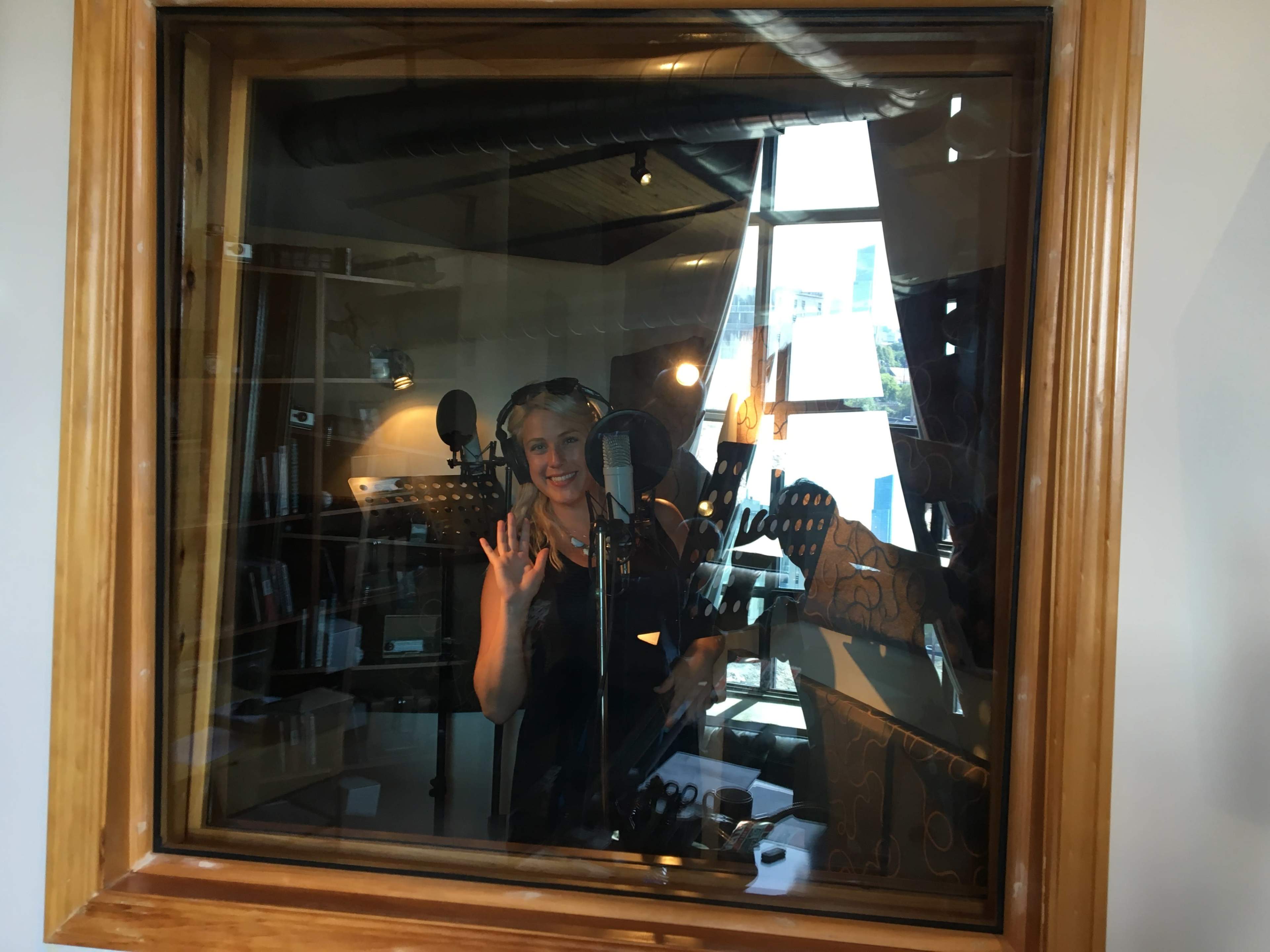 A person stands inside a sound recording studio, smiling and gesturing while microphone equipment surrounds them.