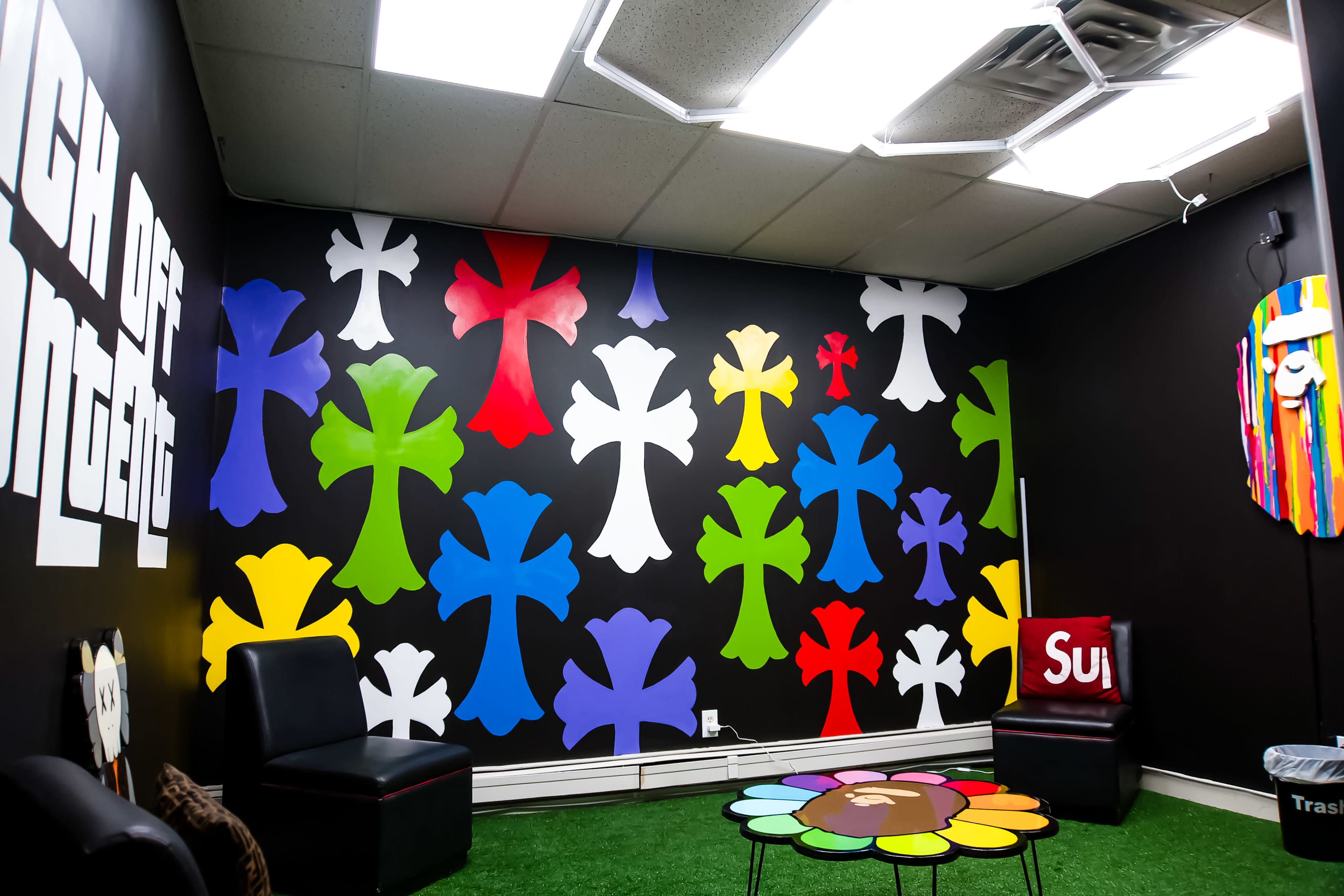 The image shows a room with a black wall covered in colorful cross-shaped decorations and a table with a flower design, accompanied by black chairs and a red object labeled "Sup."