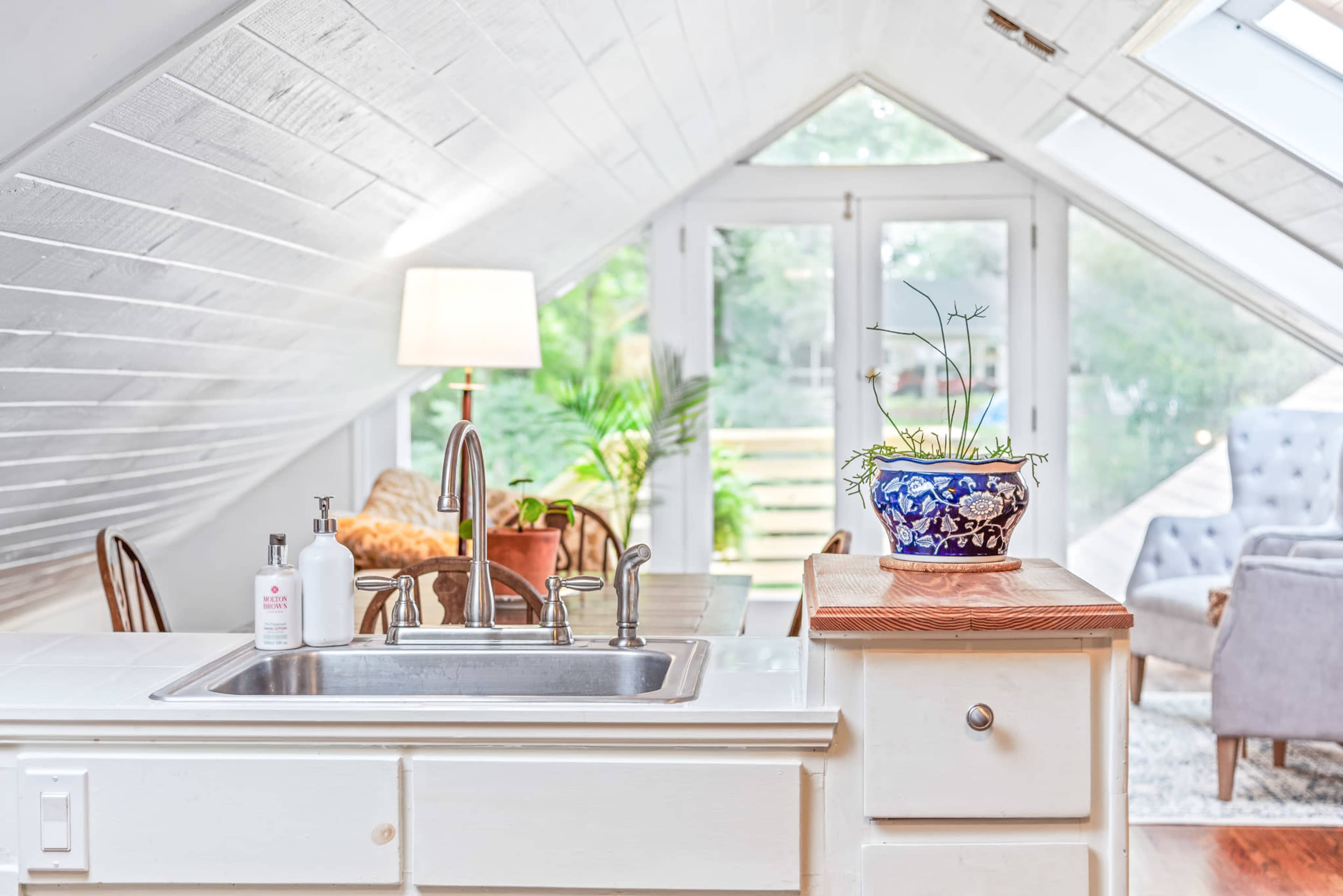 A bright, airy attic kitchen features a stainless steel sink, a decorative plant on the countertop, and large windows that bring in natural light.