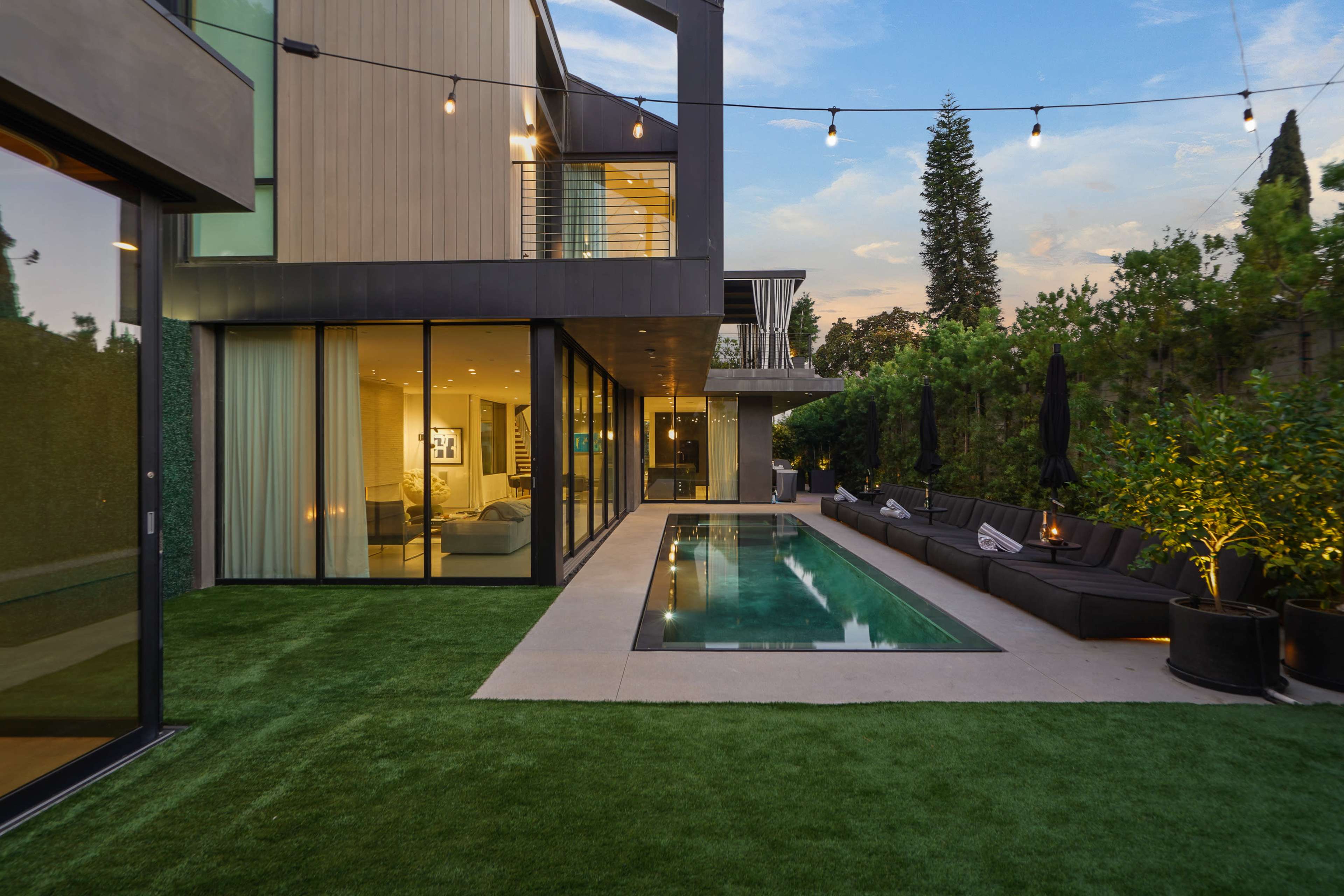 A modern home with large glass windows features a sleek pool and a landscaped yard with outdoor seating under string lights.