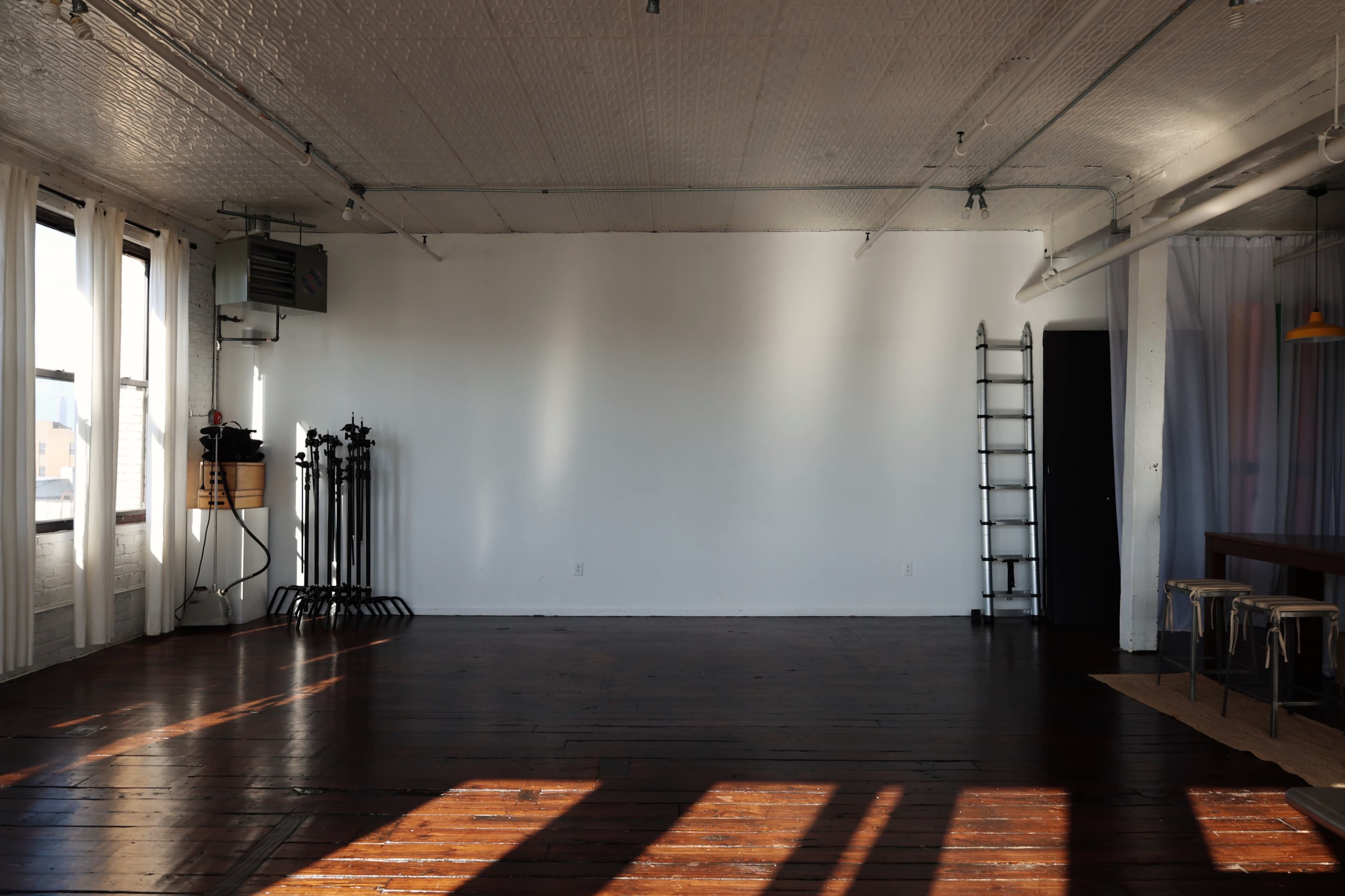 The image shows an empty room with wooden flooring, a white wall, and minimal furnishings, including a ladder and equipment stands along one side.