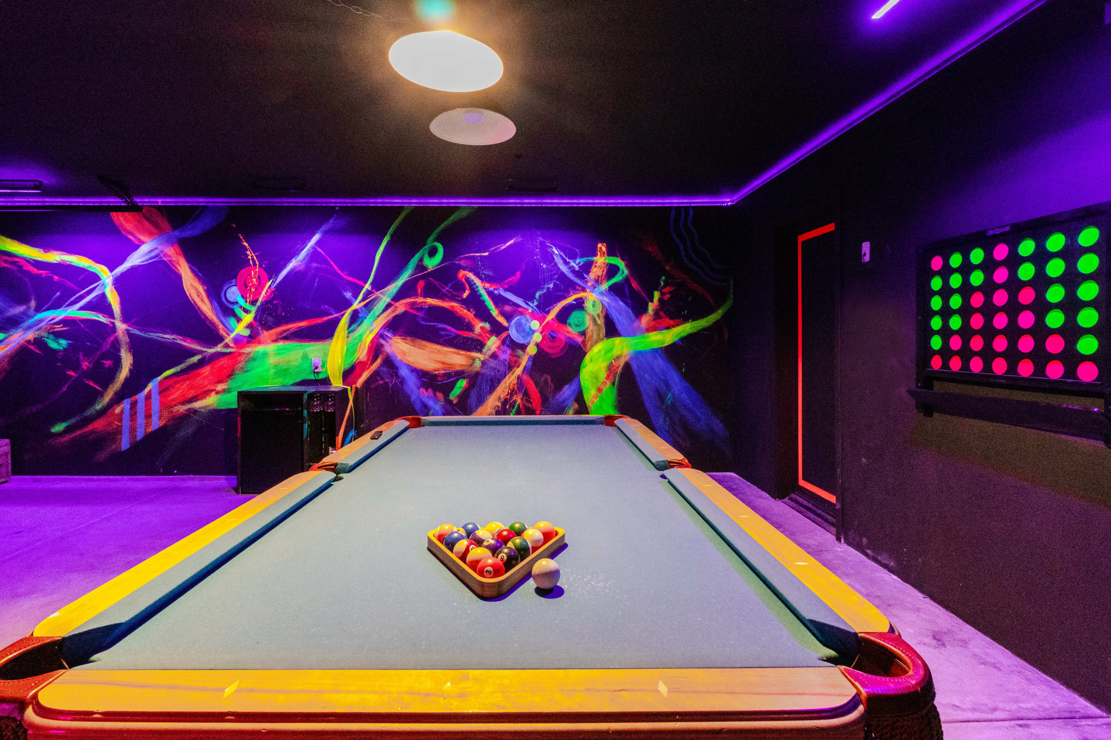A pool table set up with colored balls arranged in a triangle, surrounded by vibrant wall art and neon lighting.