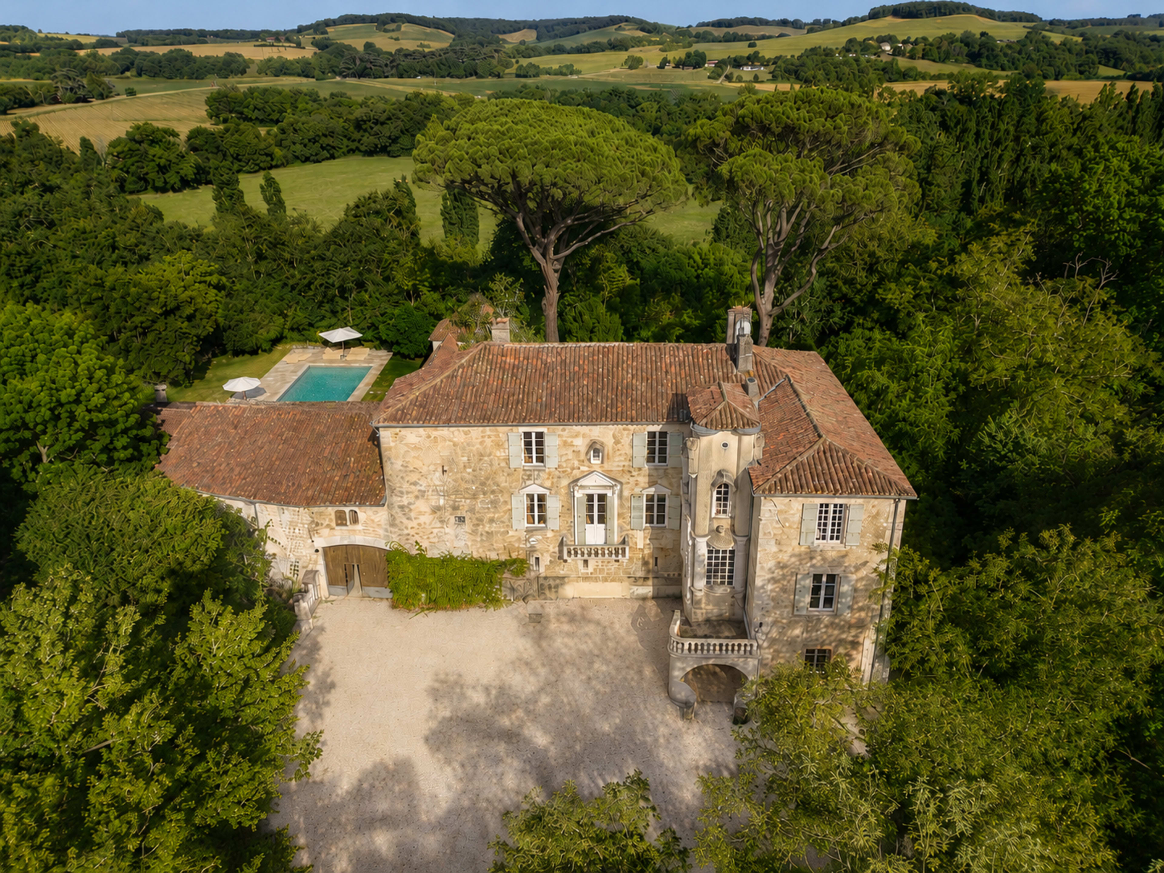 The image shows a large, stone mansion surrounded by lush greenery and rolling hills, with a swimming pool visible in the backyard.