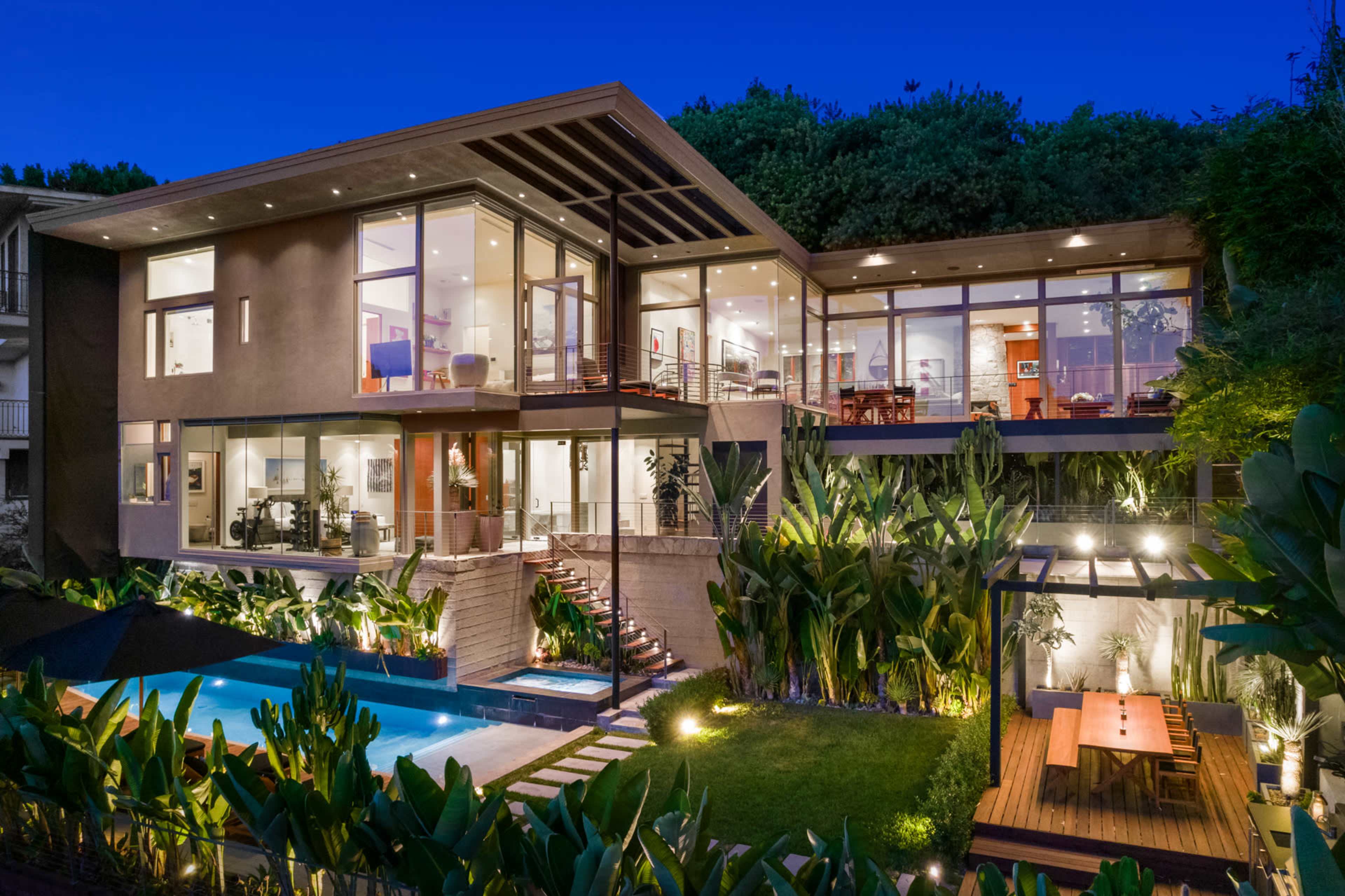 The image shows a modern house with large glass windows, a swimming pool, and landscaped greenery, illuminated at dusk.