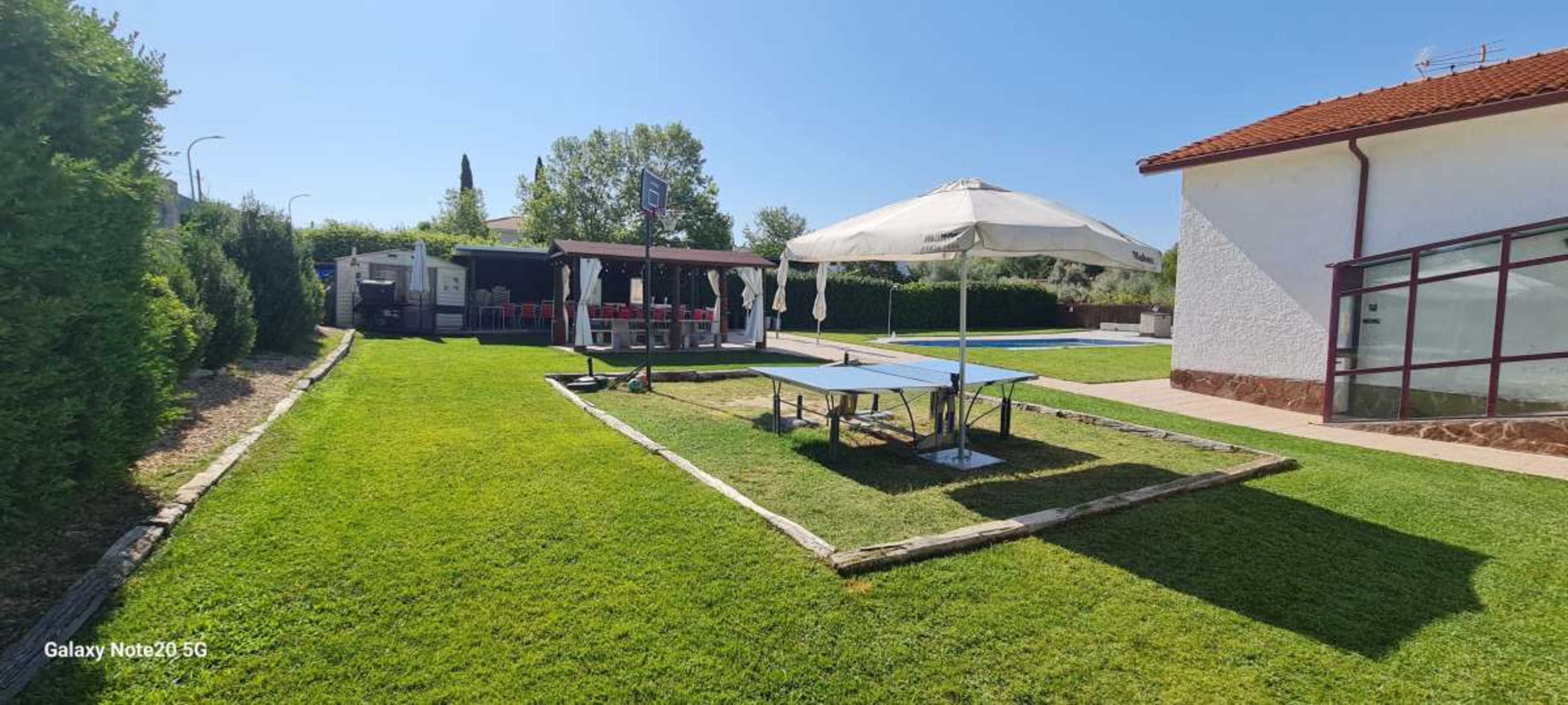 The image shows a well-maintained garden area with a ping pong table, a shaded seating area in the background, and a swimming pool on the right side.