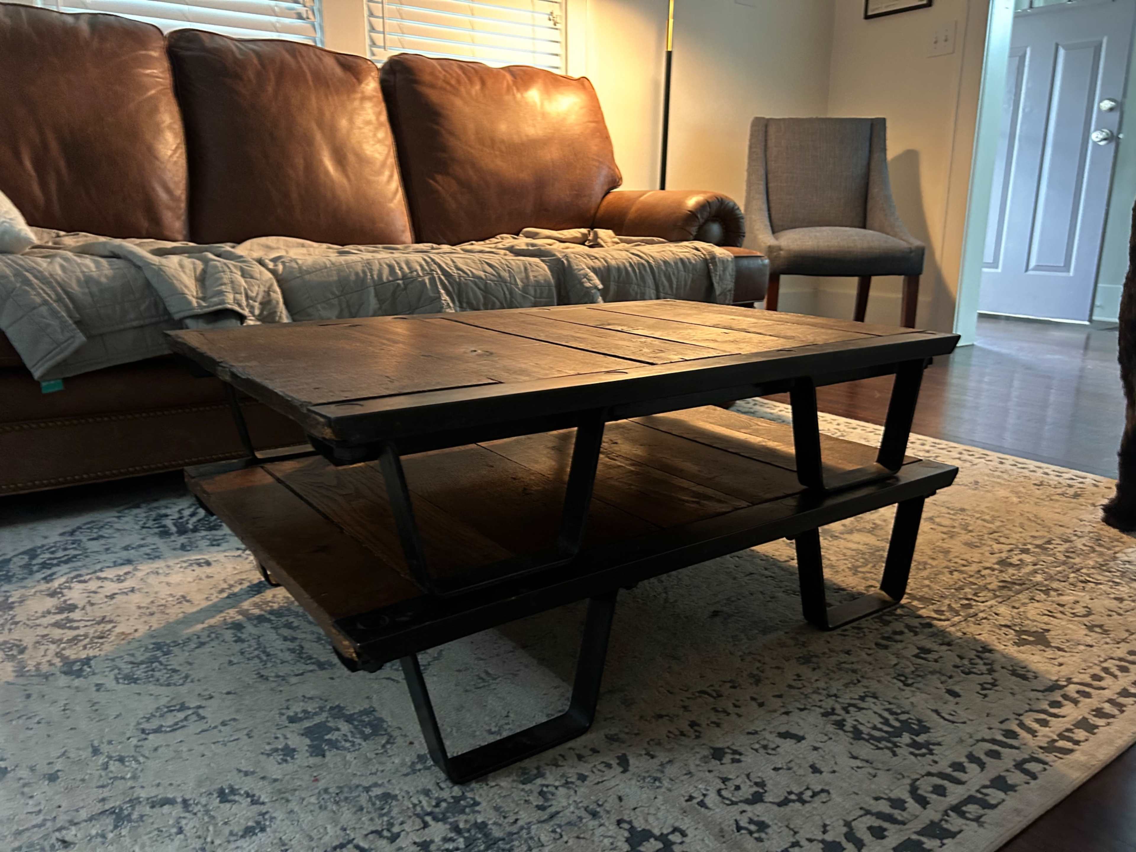 The image shows a wooden coffee table with a stacked design, placed in a living room featuring a brown leather sofa and a blue chair.