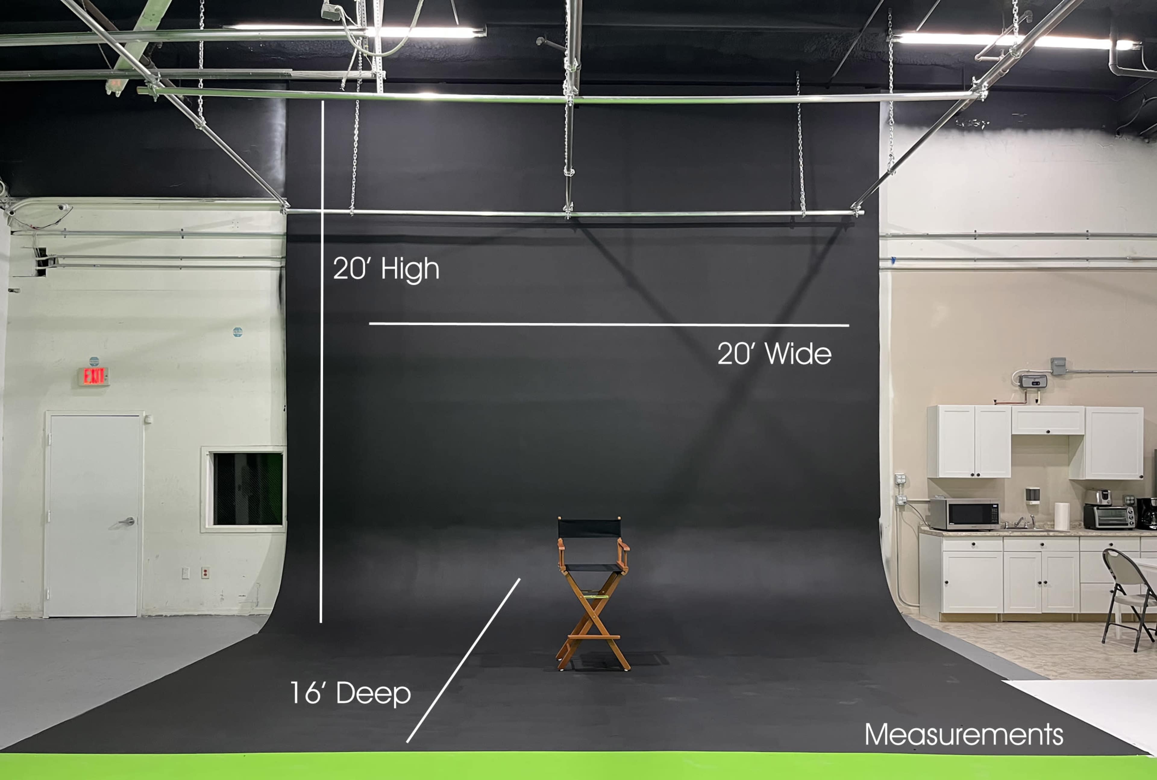 A photography studio with a black backdrop measuring 20 feet high and 20 feet wide, featuring a director's chair positioned in front and a kitchenette area visible to the side.