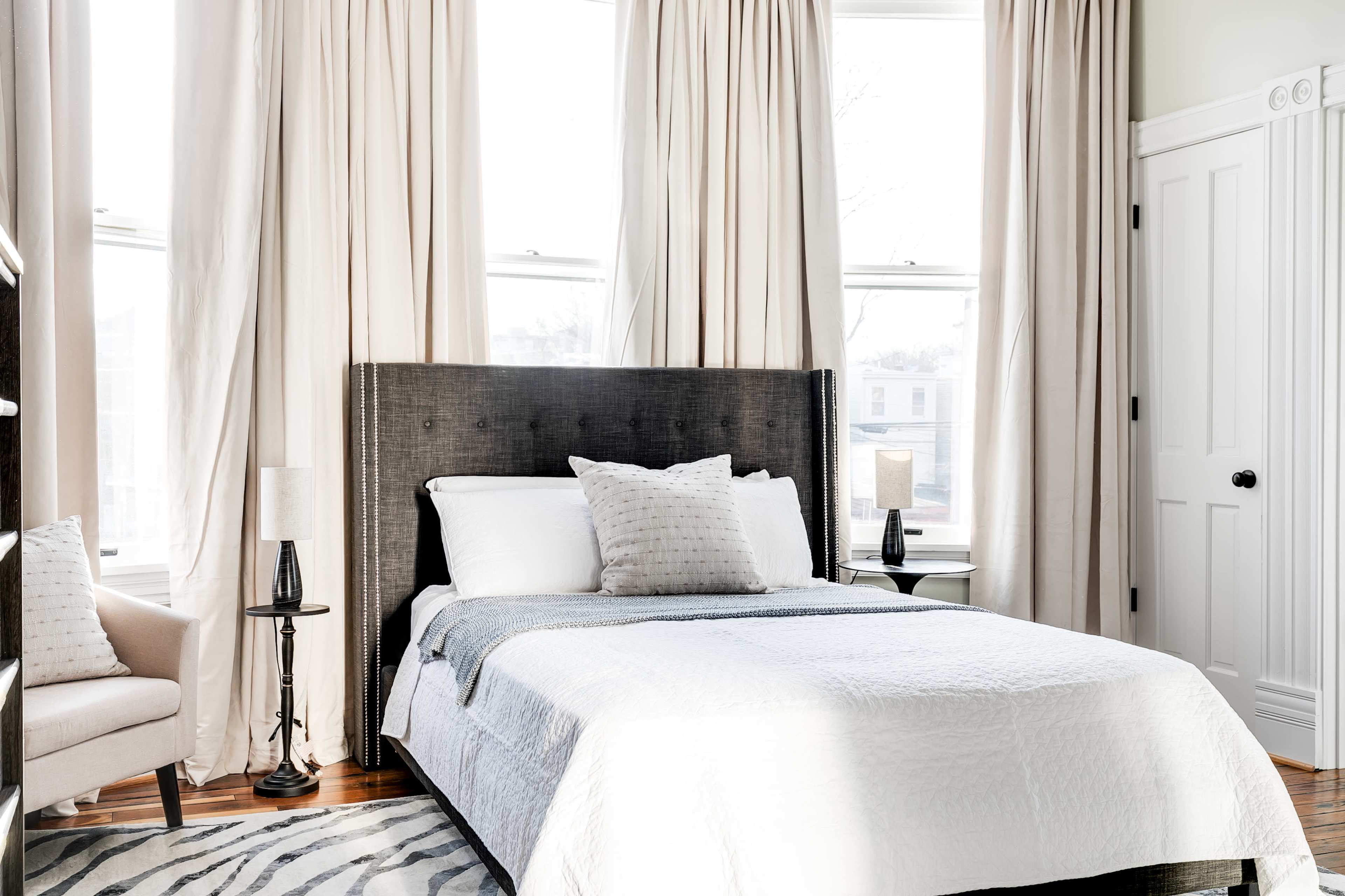 The bedroom features a bed with a dark headboard, adorned with a white quilt and gray throw, surrounded by large windows dressed with light curtains.