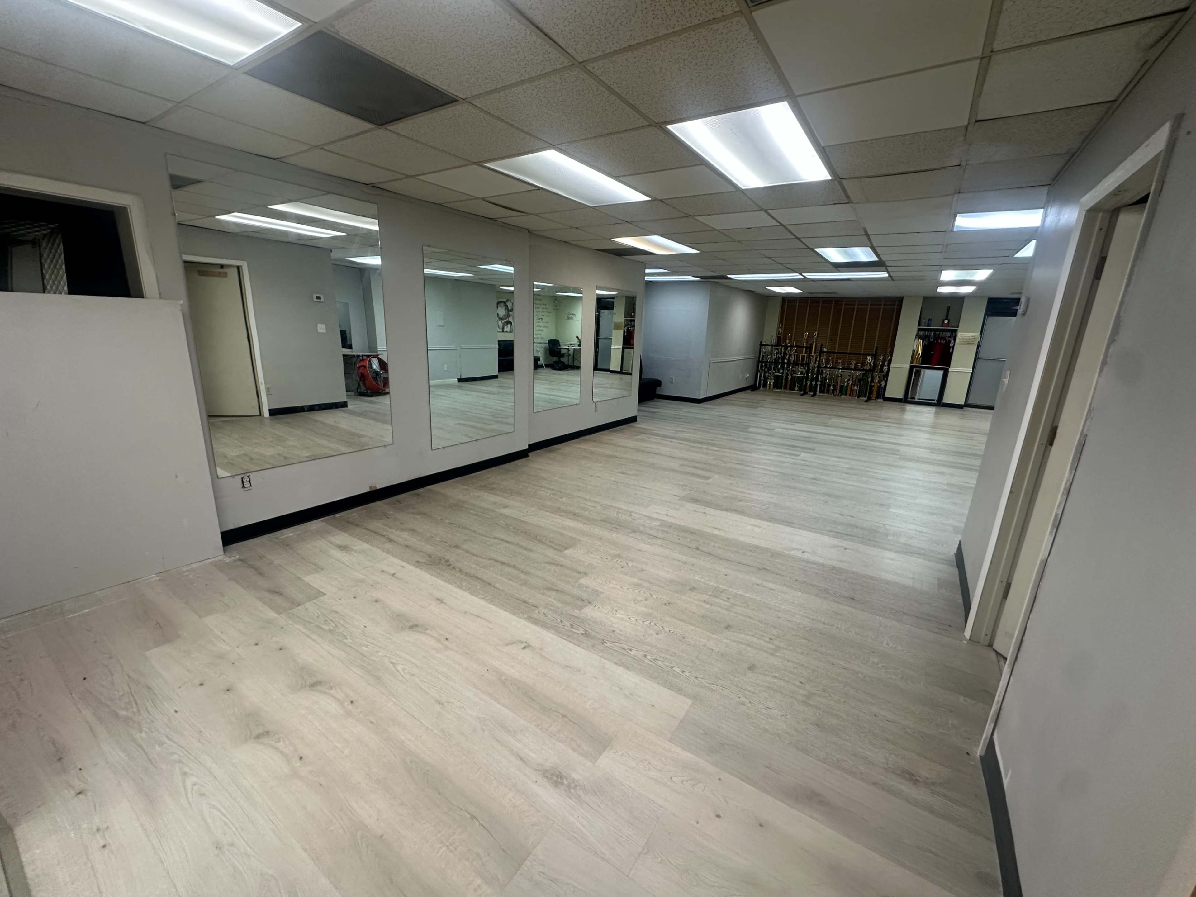 The image shows a spacious, empty room with light-colored flooring, mirrored walls, and fluorescent ceiling lights.