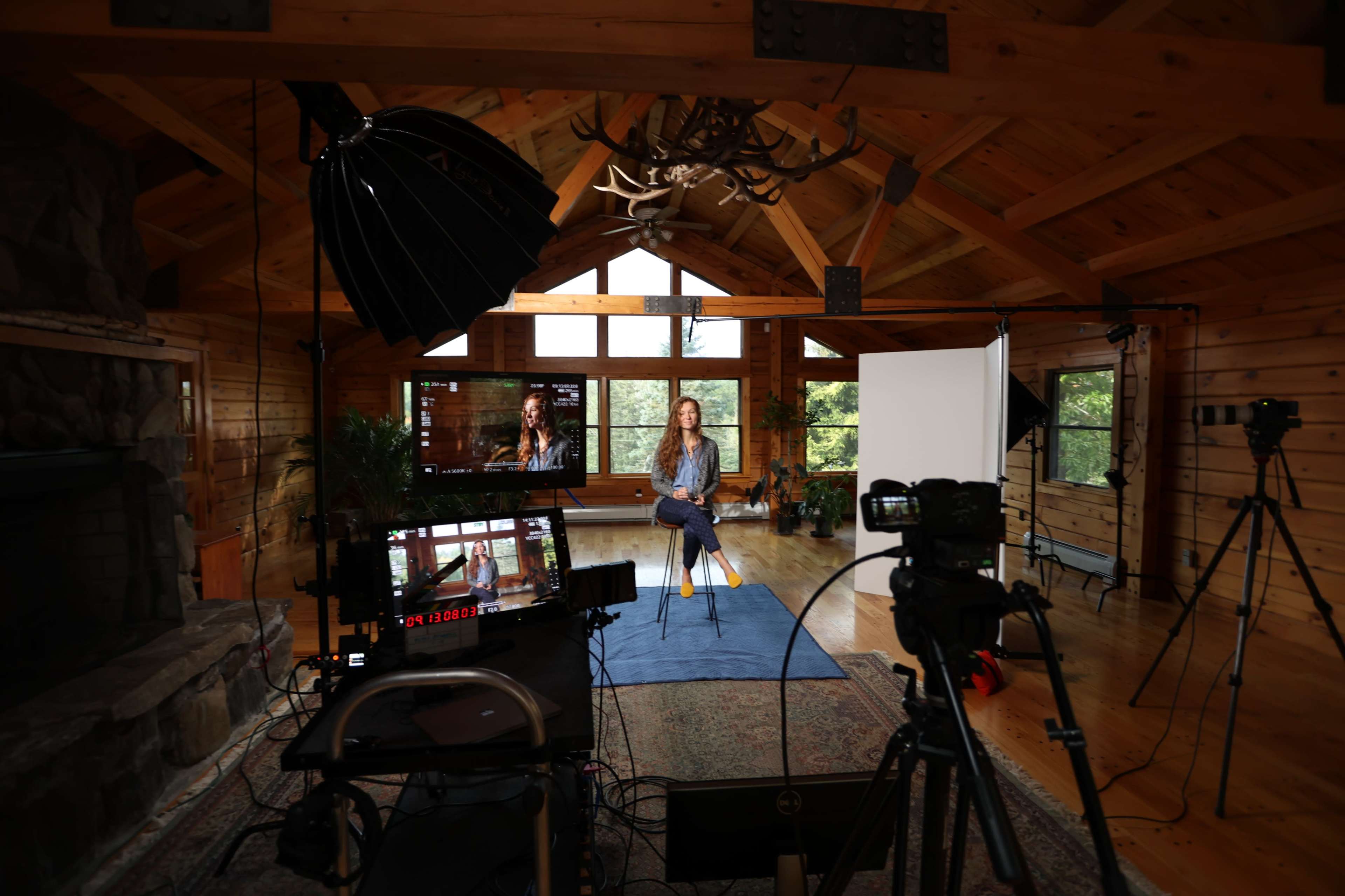 A woman sits on a stool in a wooden room set up for filming, surrounded by cameras and lighting equipment.