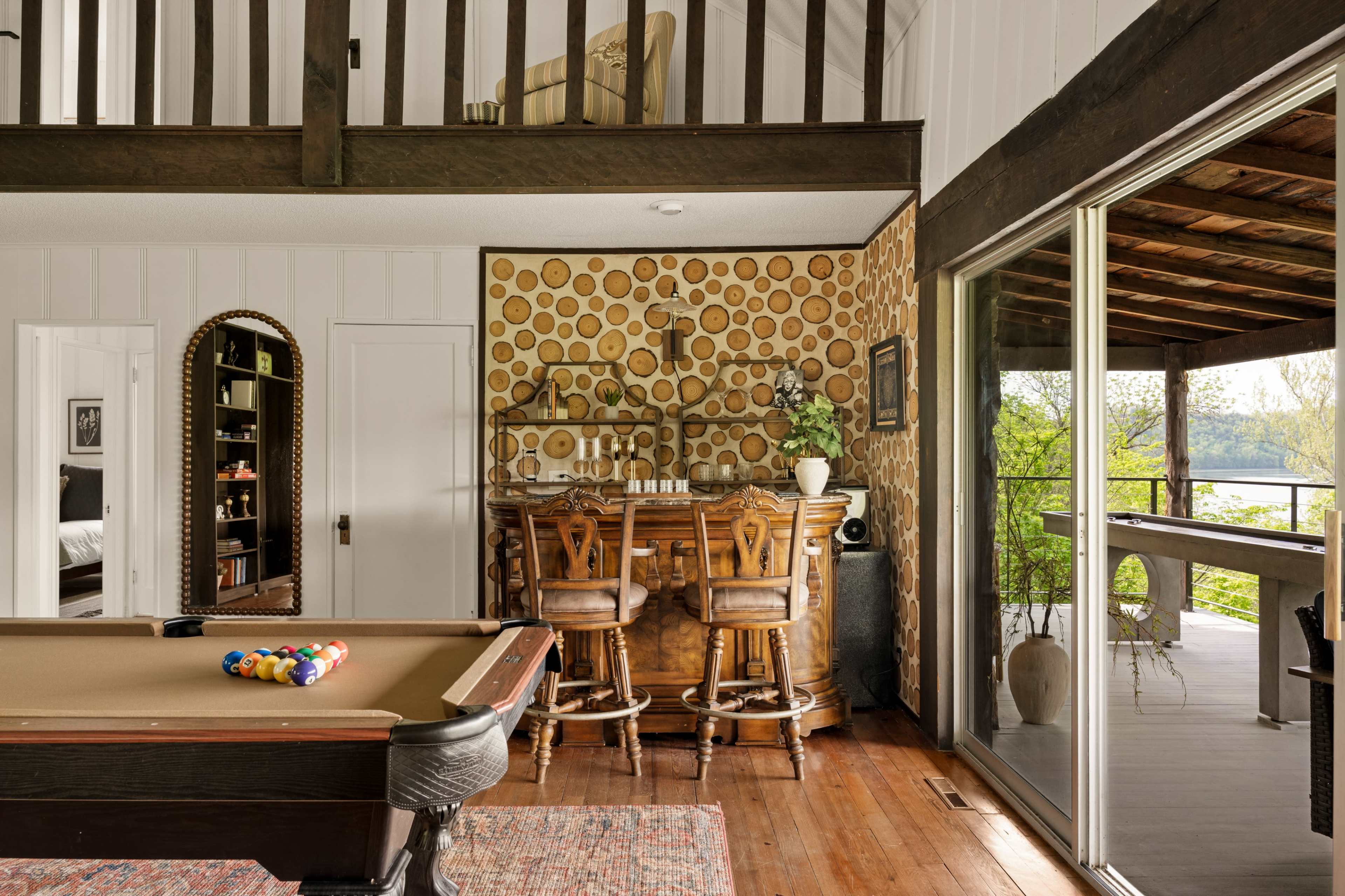 The image shows a rustic game room with a billiard table, a wooden bar with stools, and a sliding glass door leading to an outdoor area with a view.