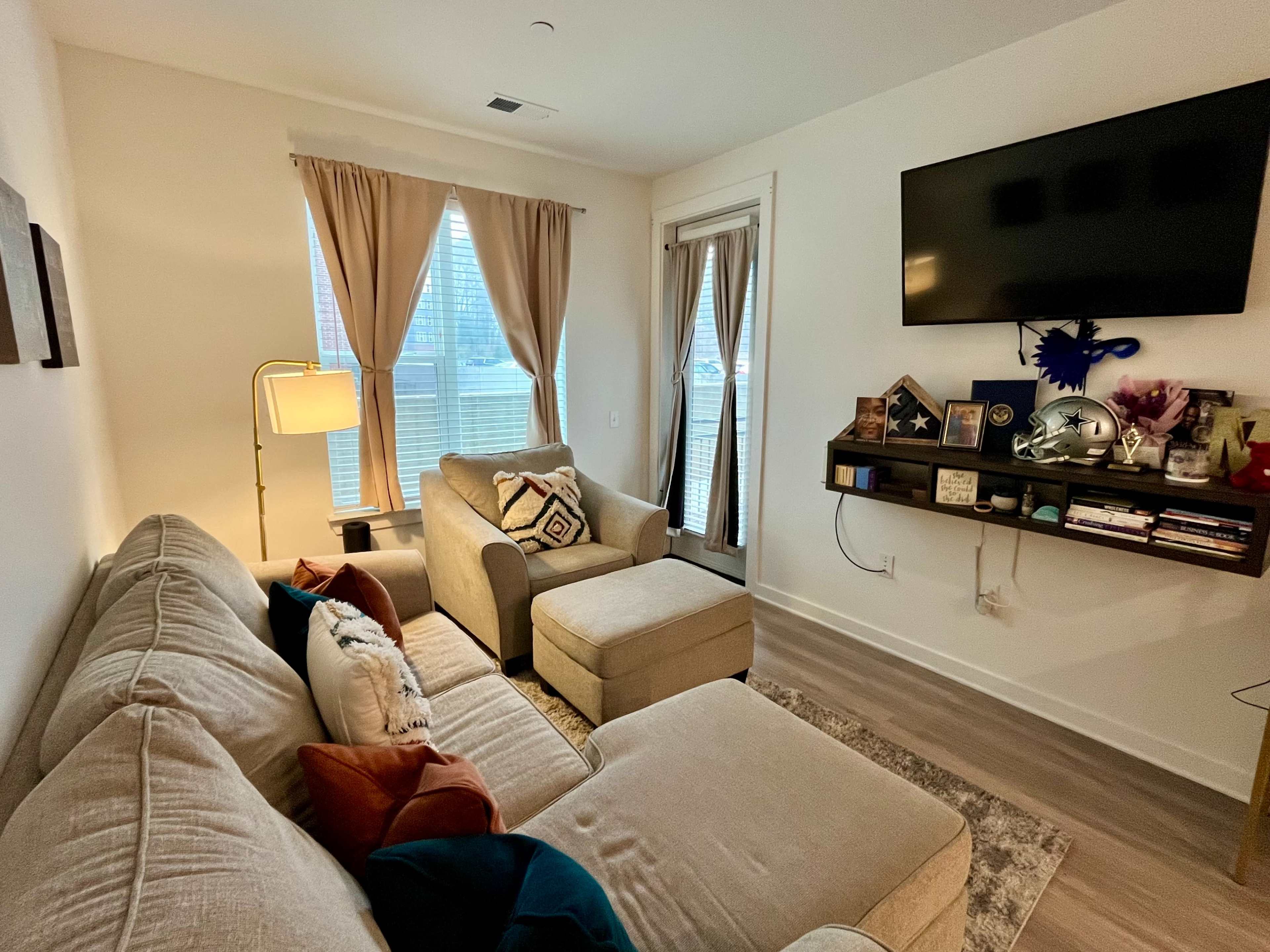 The image shows a cozy living room with a beige sectional sofa, a small armchair, a floor lamp, and a mounted flat-screen TV on the wall.