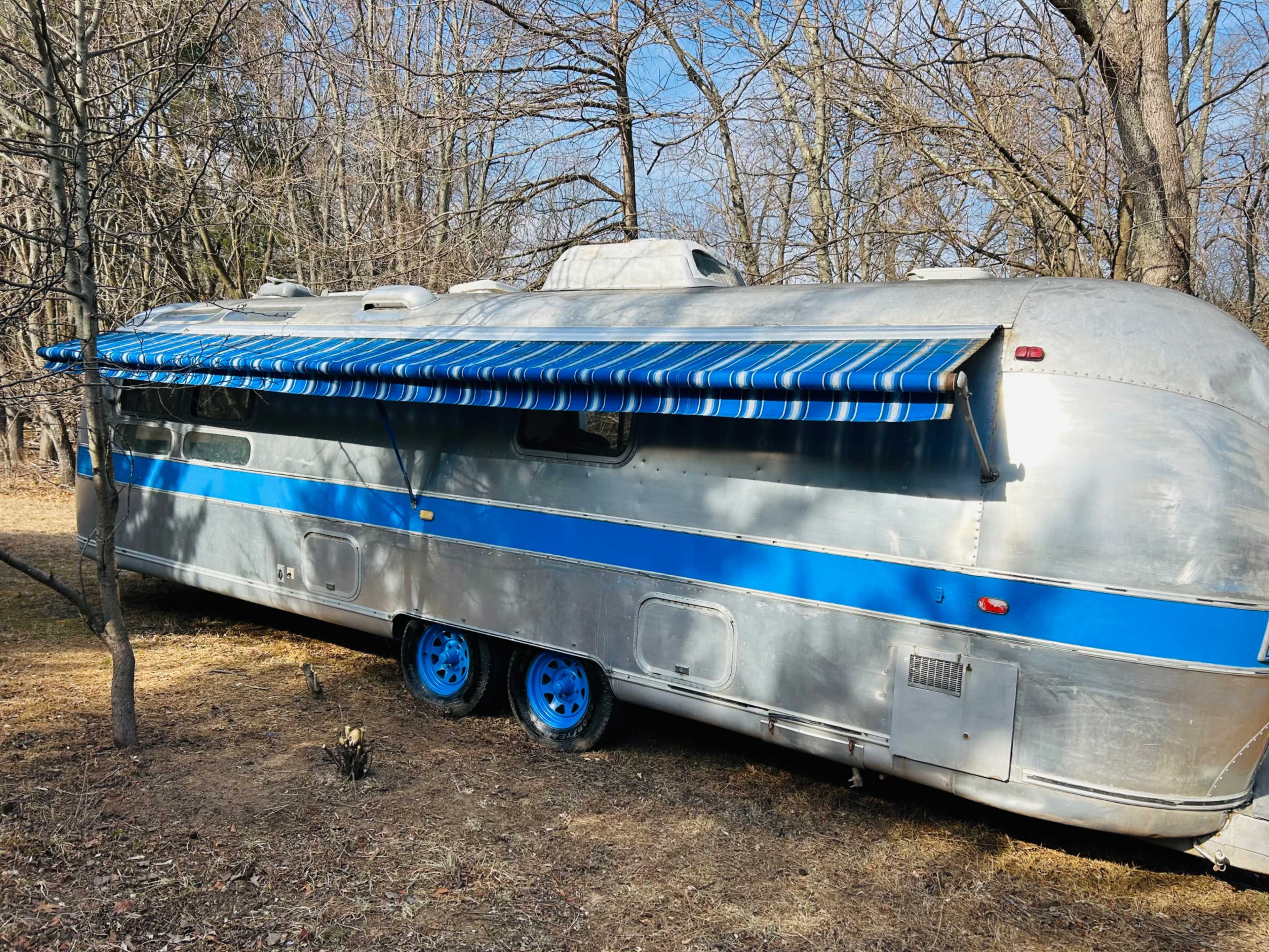 The image shows a vintage silver Airstream trailer parked in a wooded area with a blue awning extended.