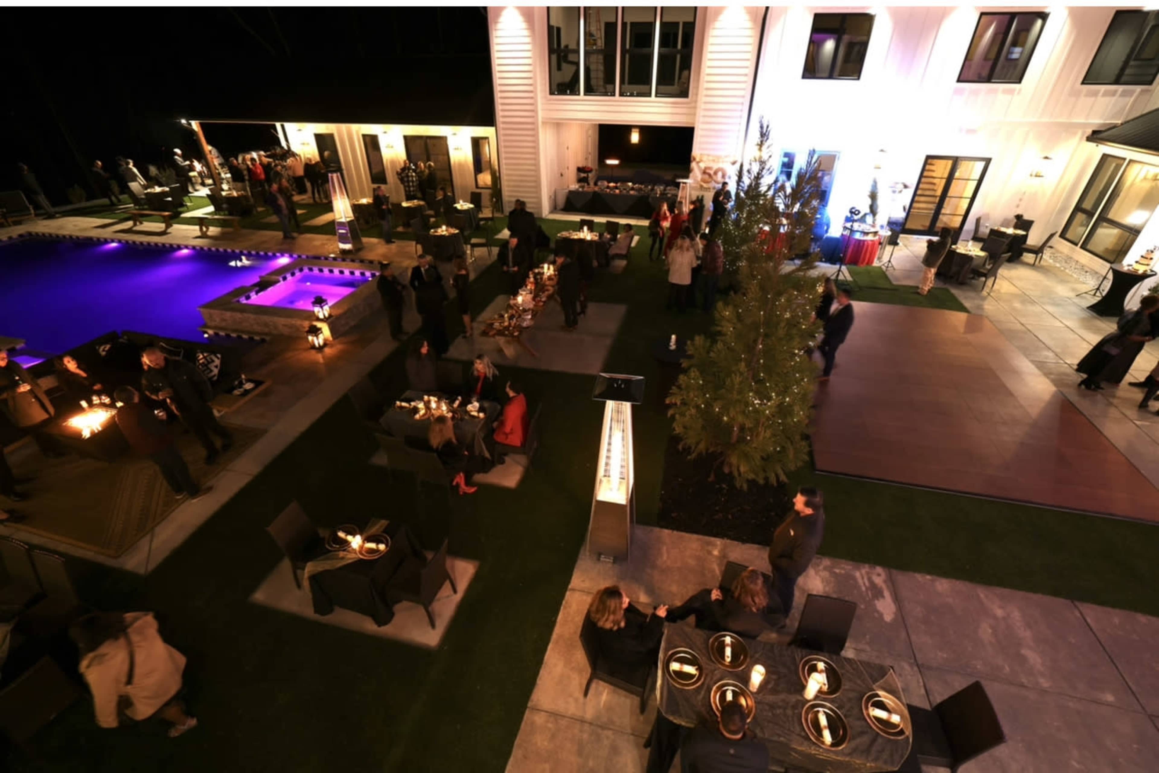 The scene shows a nighttime outdoor event at a modern home, featuring a pool illuminated with purple lighting and guests mingling around tables and fire pits.
