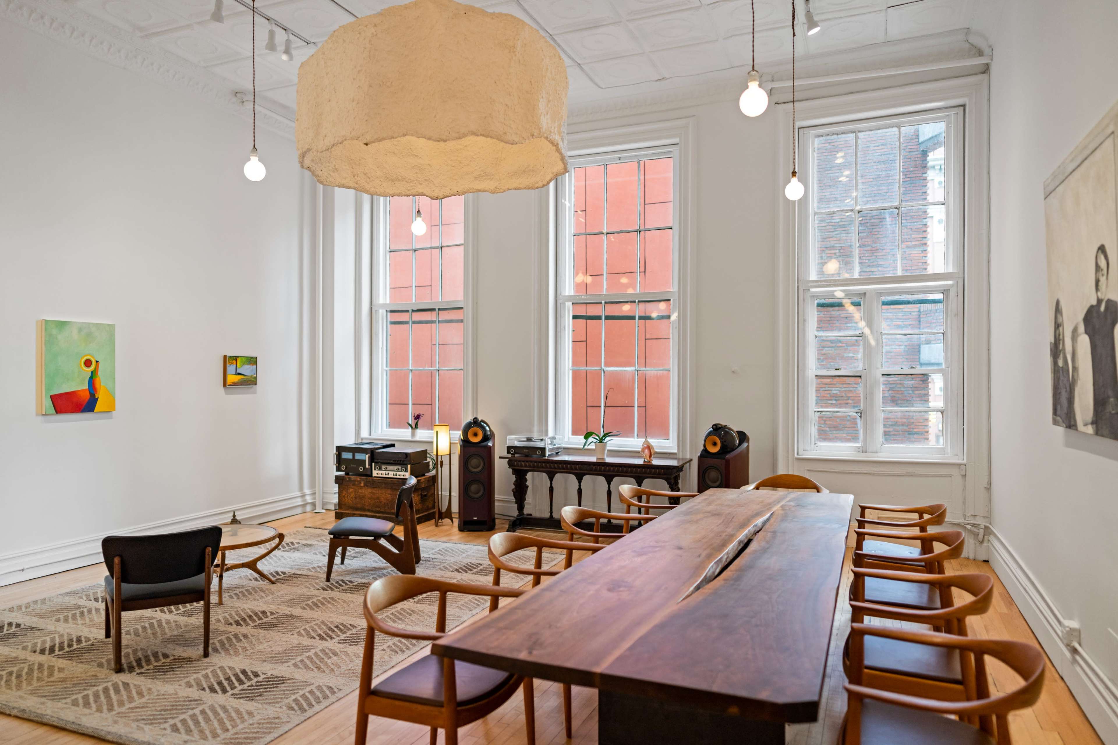 A spacious room features large windows, a long wooden dining table surrounded by chairs, and a seating area with an armchair and bookshelf.