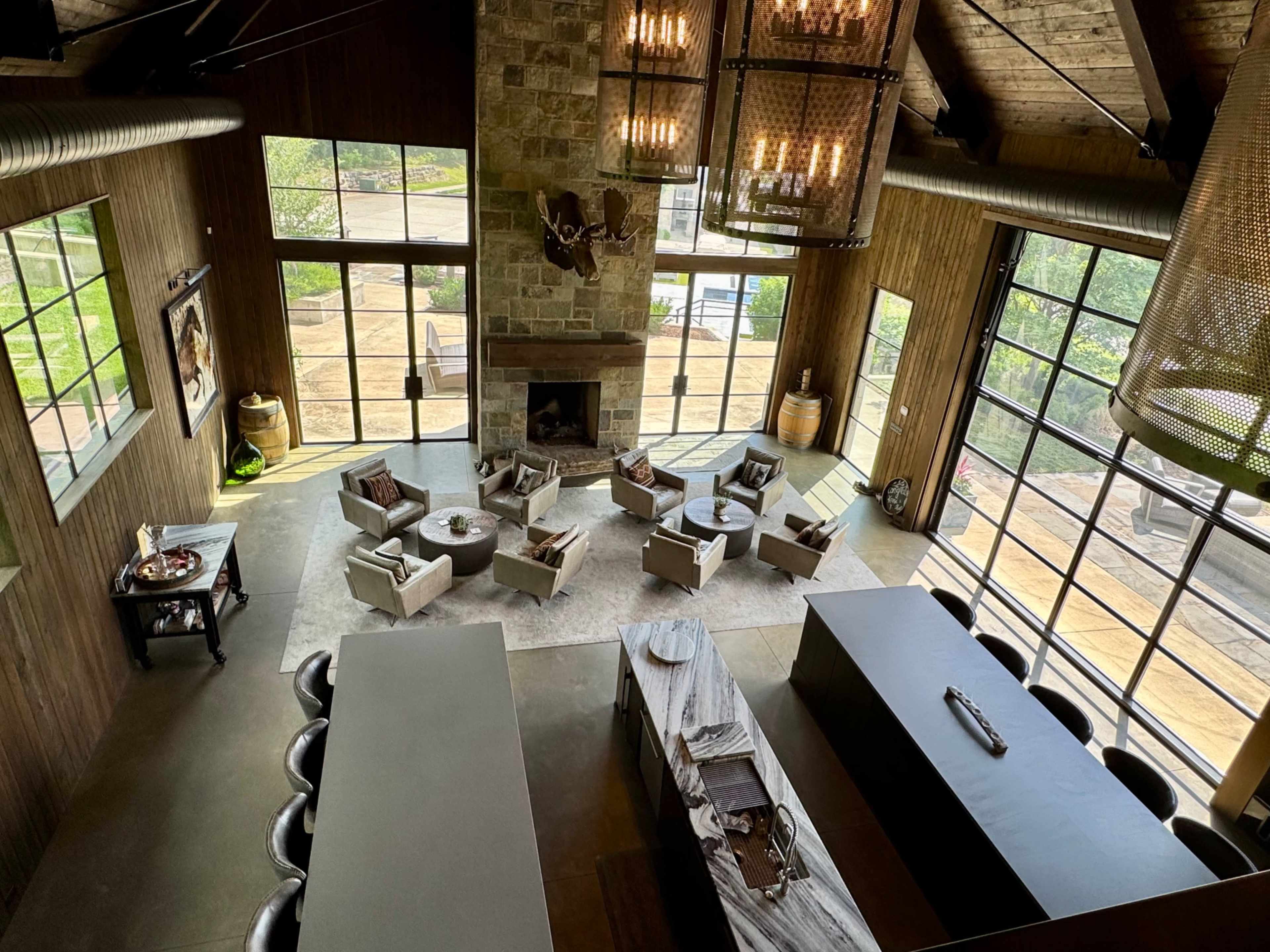 The image shows a spacious, modern living area with large windows, a stone fireplace, and a blending of indoor and outdoor spaces featuring seating arrangements and a long dining table.