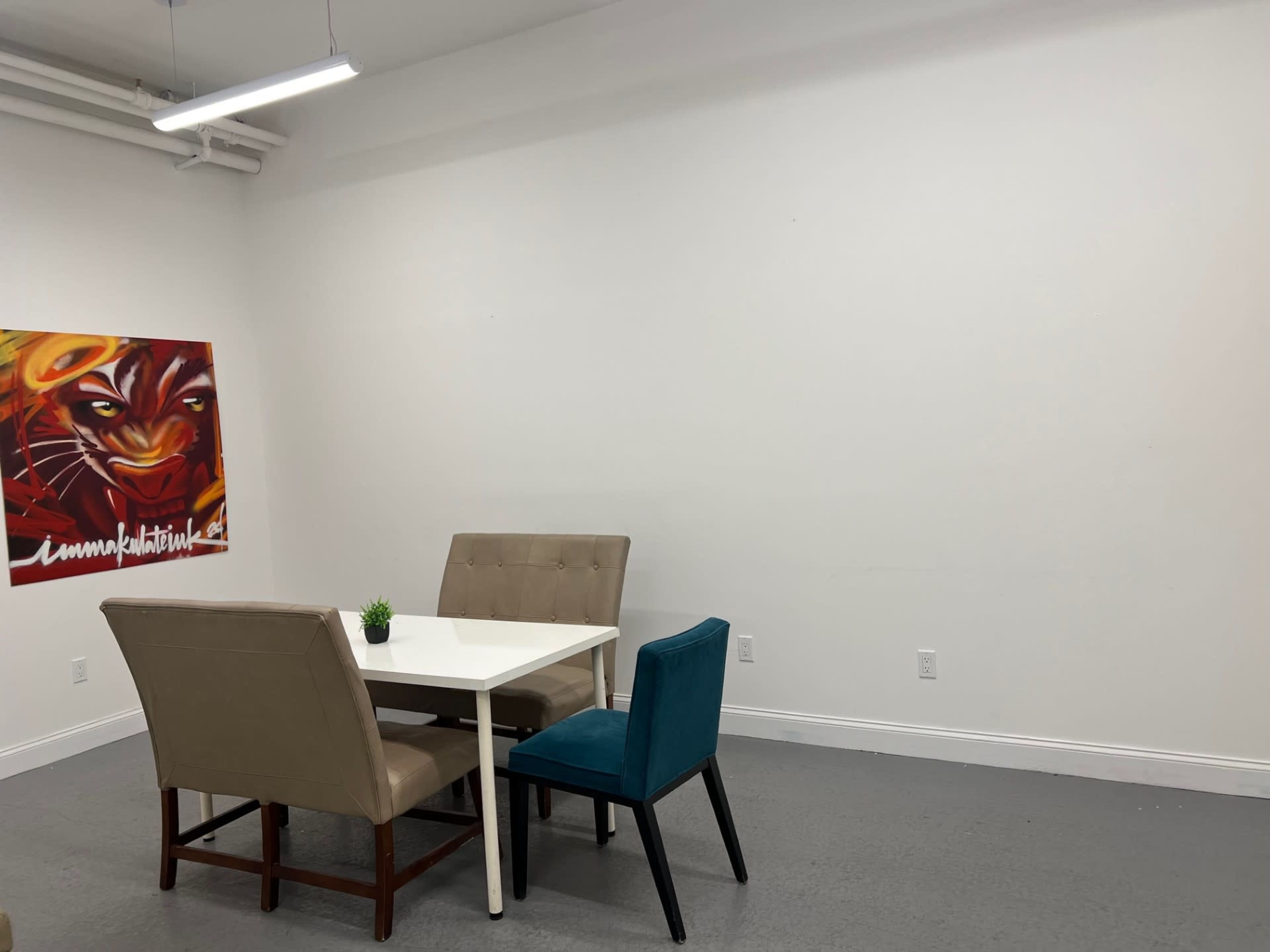The image shows a small dining area with a white table surrounded by four chairs in a minimalistic room with white walls and a colorful mural on the left.