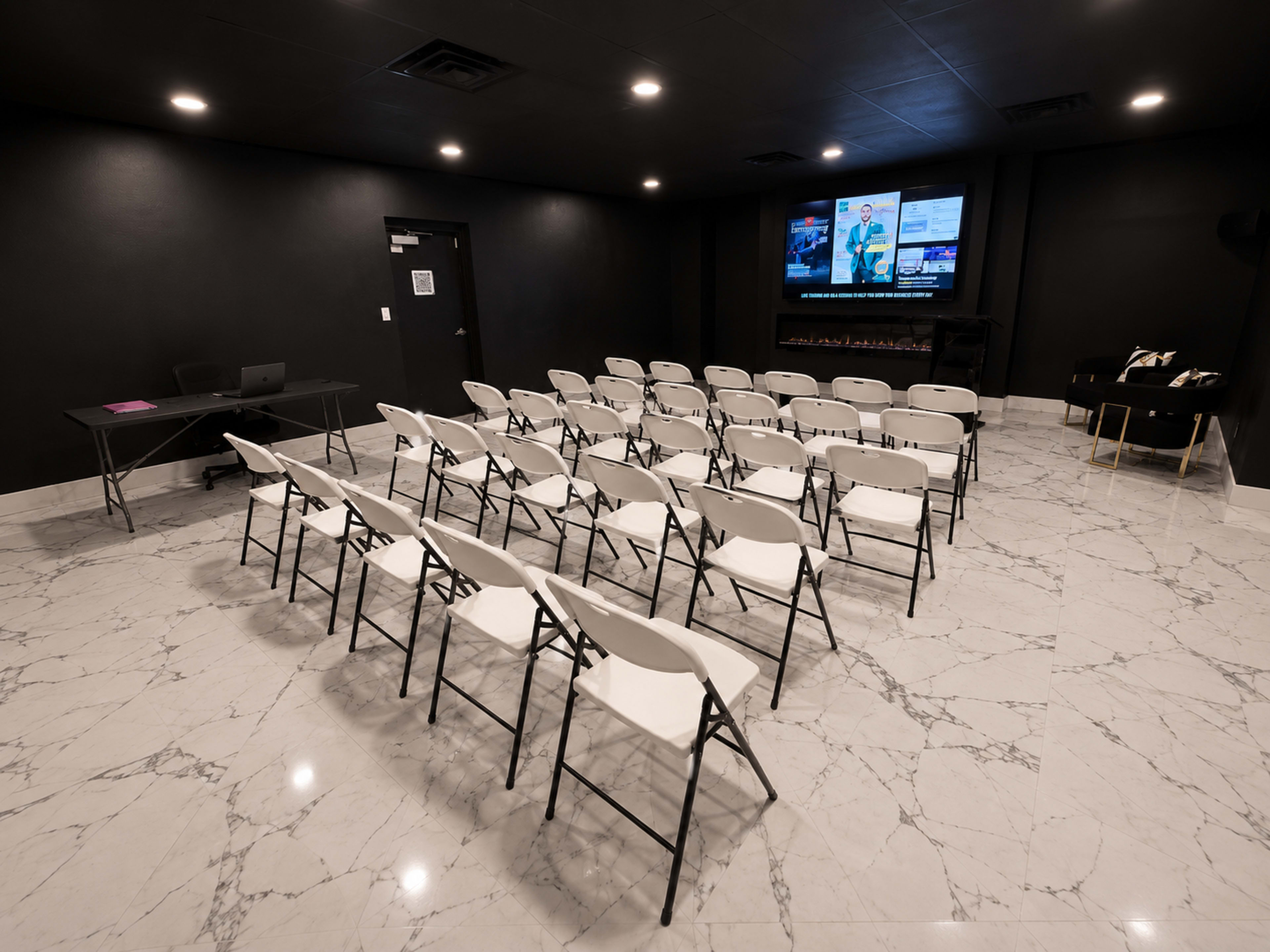 The image depicts a minimalistic conference room with rows of white folding chairs facing dual screens on a black wall, situated on a marble floor.