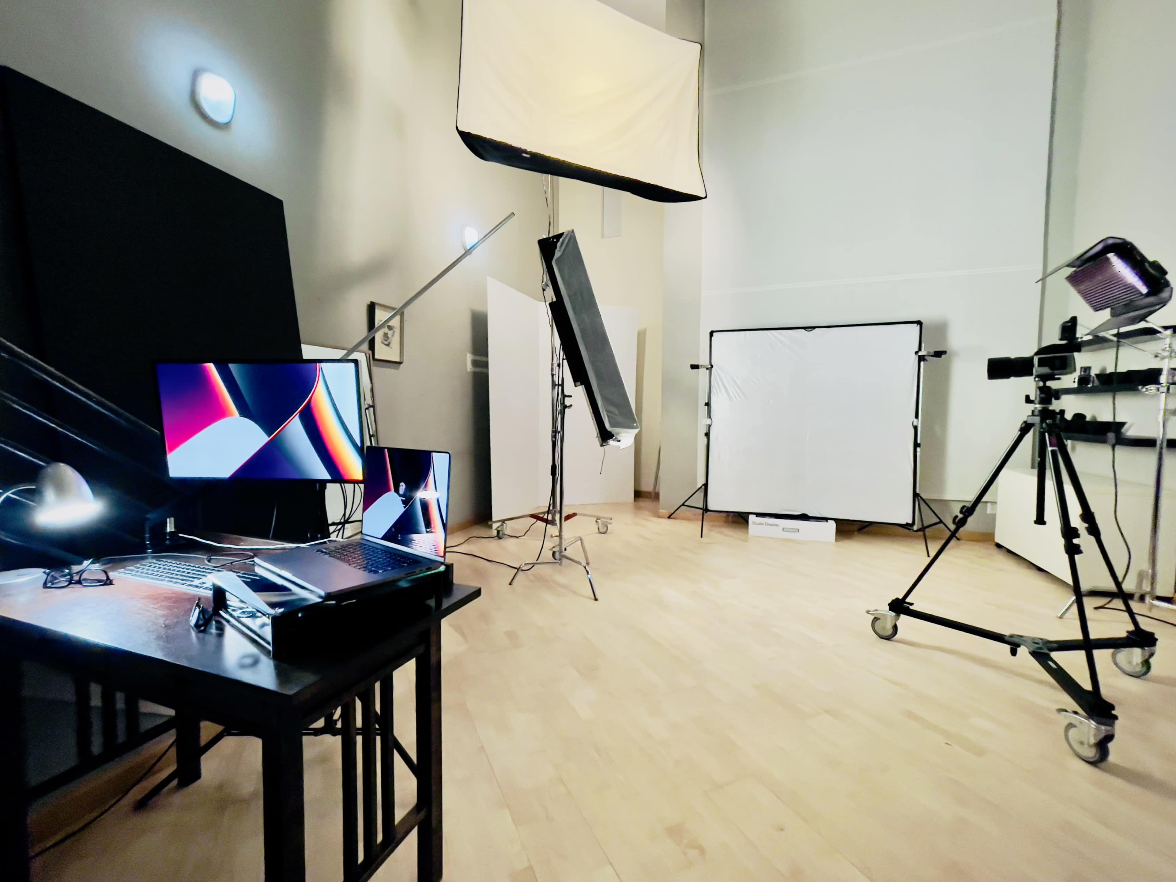 A well-lit studio setup features a table with a laptop and external monitor, along with various lighting equipment and backdrops arranged around the space.