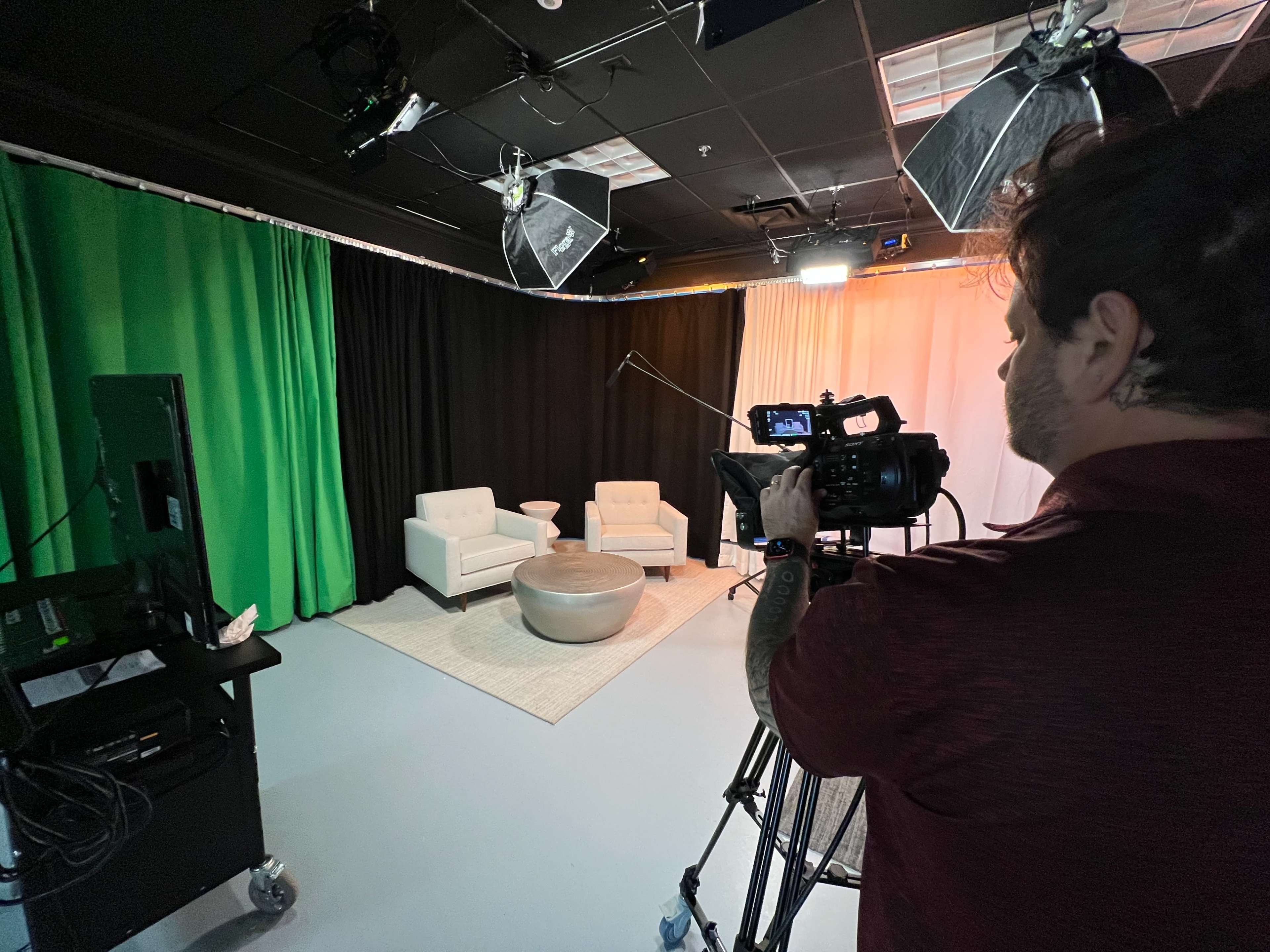 A person operates a camera in a television studio setting featuring two white chairs and a round table against a backdrop of colored curtains.