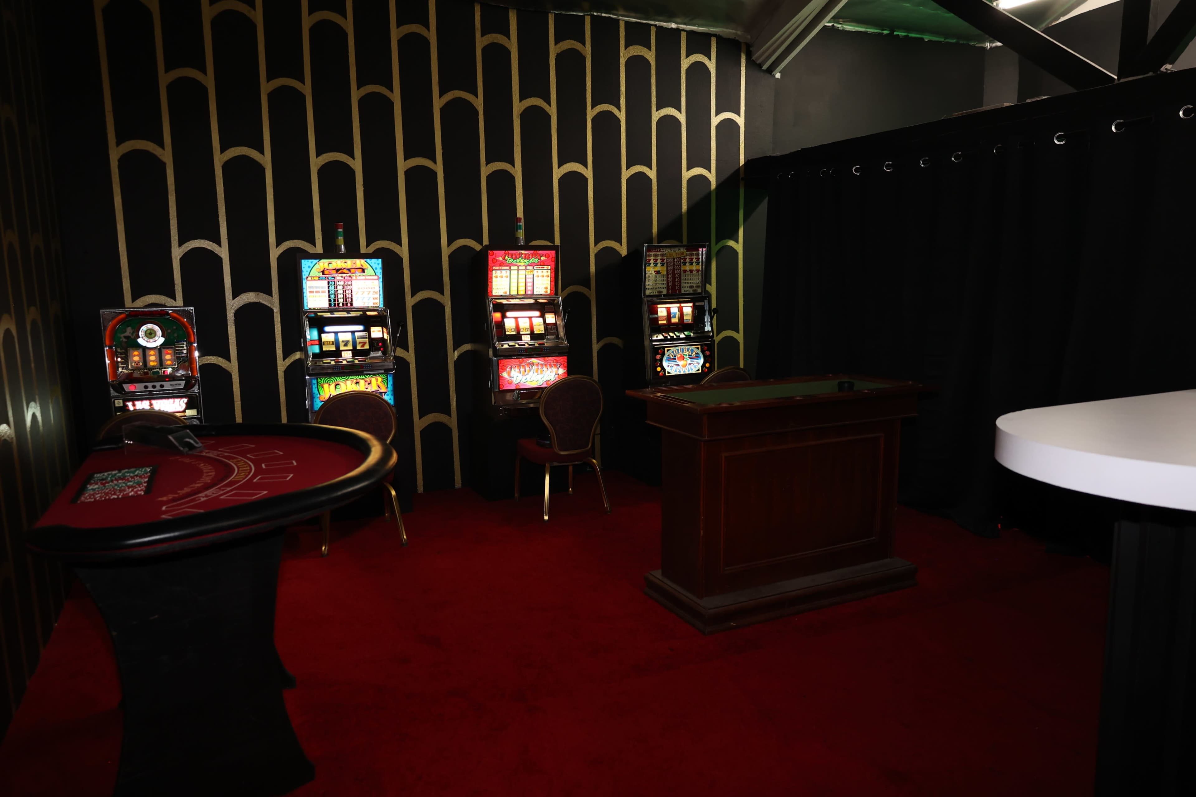 A dimly lit gambling area with a red carpet, a poker table, and several slot machines against a patterned black wall.