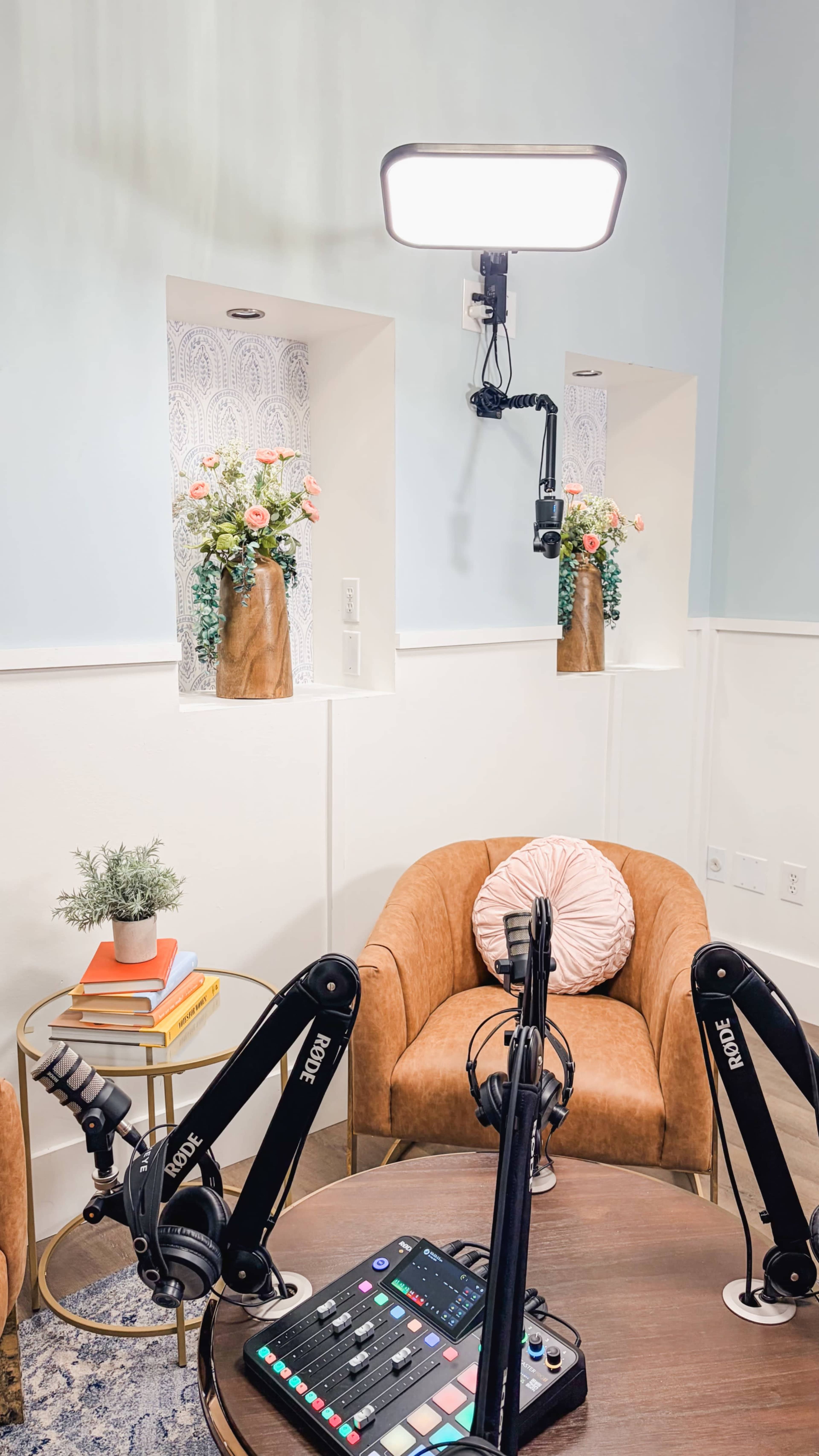 The image shows a cozy podcasting setup featuring a round wooden table, orange armchairs, and decorative plants displayed in wall niches.