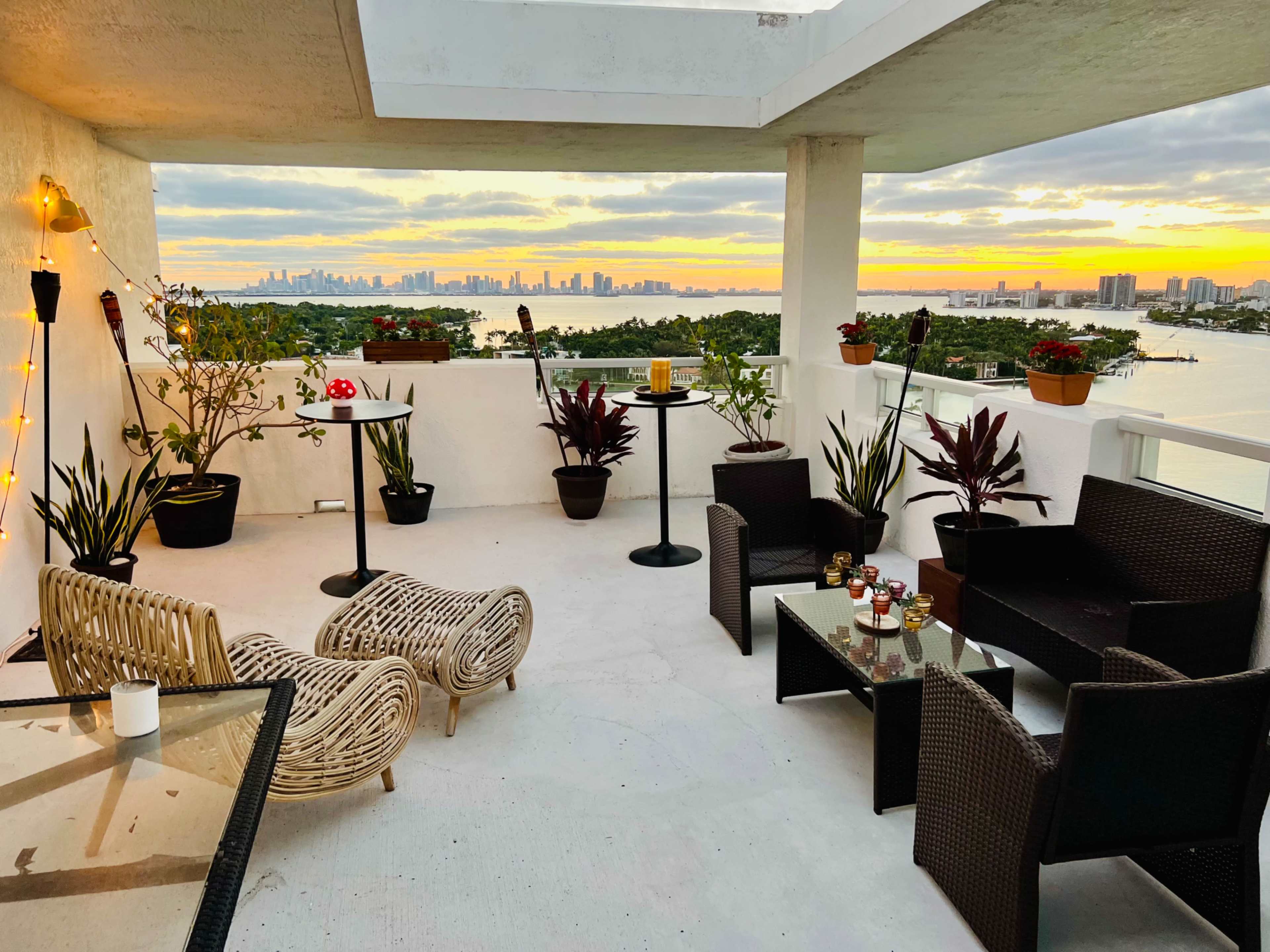 A rooftop terrace features comfortable seating arrangements, potted plants, and a panoramic view of a city skyline at sunset.