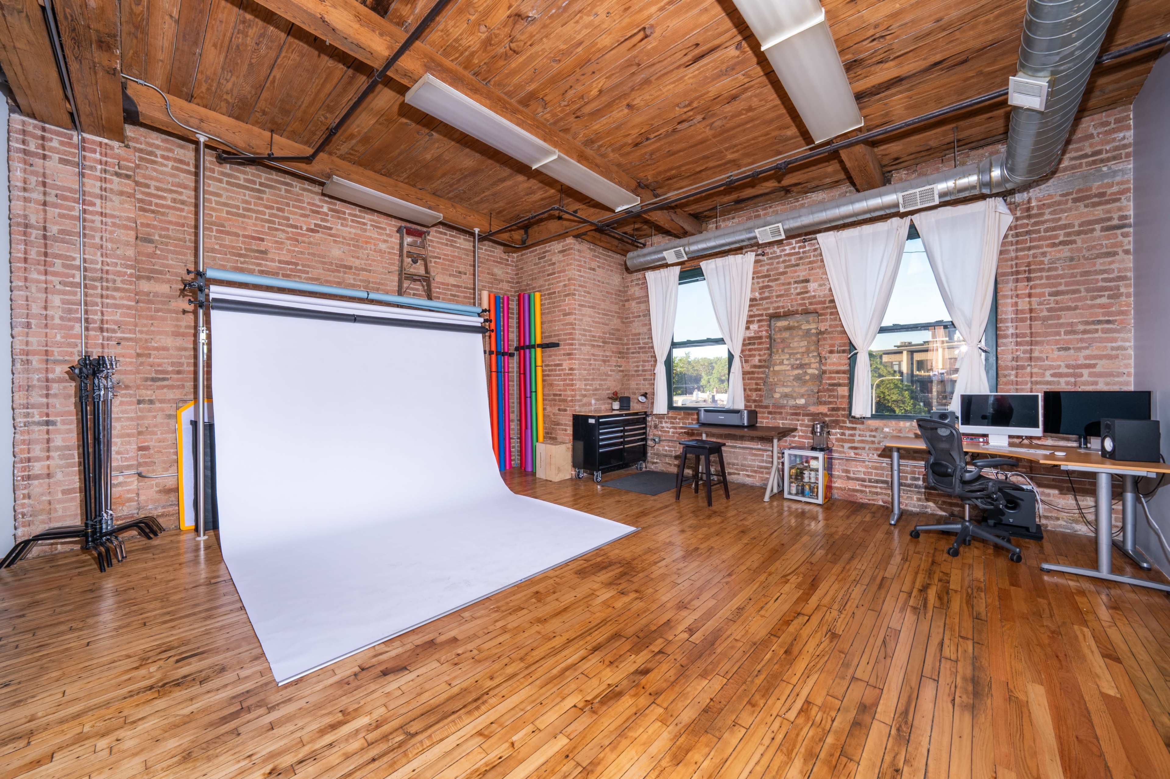 The image shows a spacious photo studio with a white backdrop, wooden flooring, large windows, and a workspace featuring a desk and computer setup.