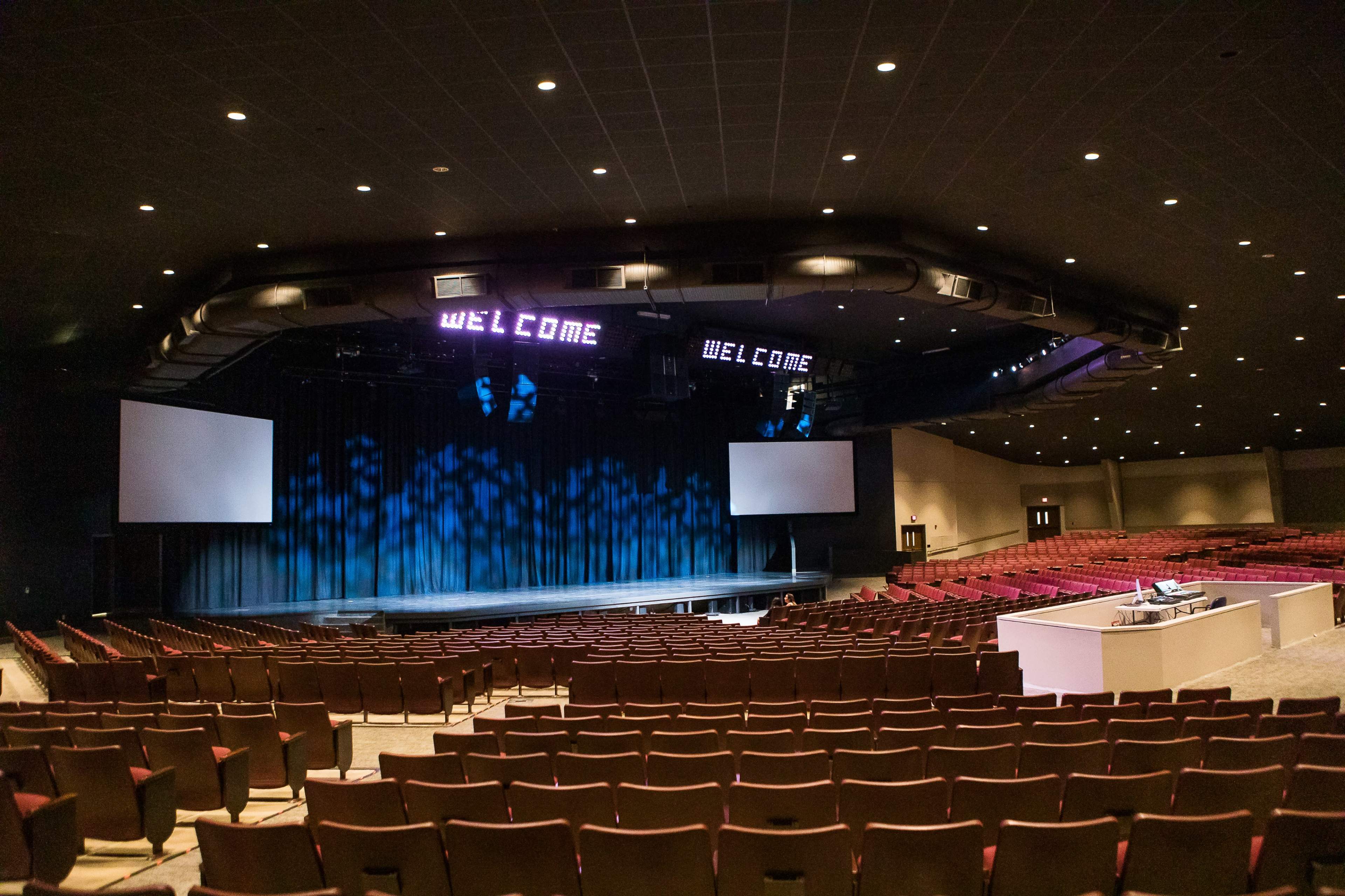 A spacious auditorium with a dark stage, a large blue curtain, and two signs that say "WELCOME" illuminated above the stage.