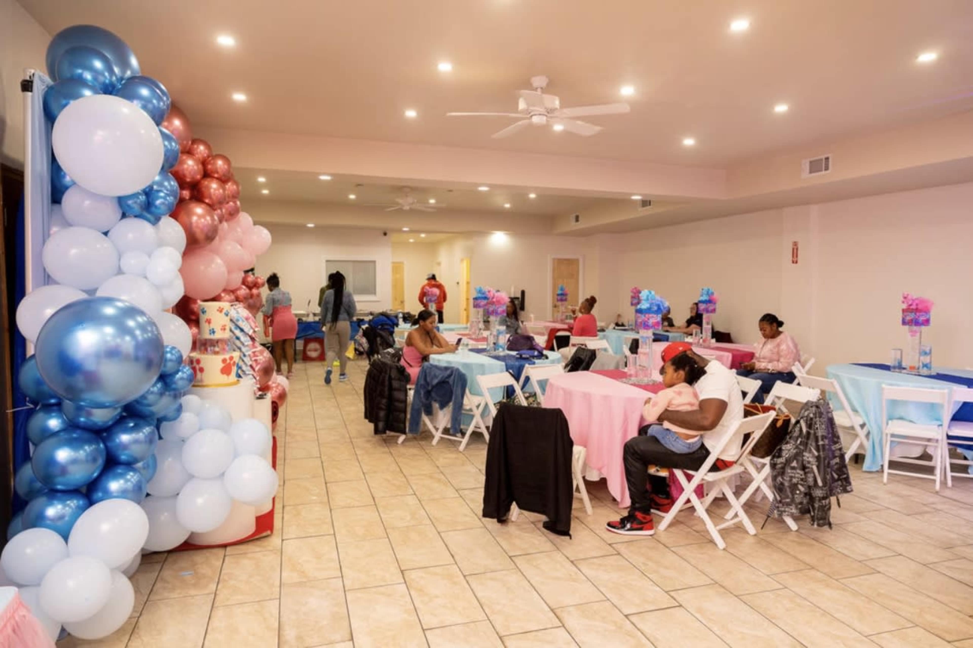 A decorated event space with tables set in pastel colors and a balloon arch in one corner, while guests engage in various activities.