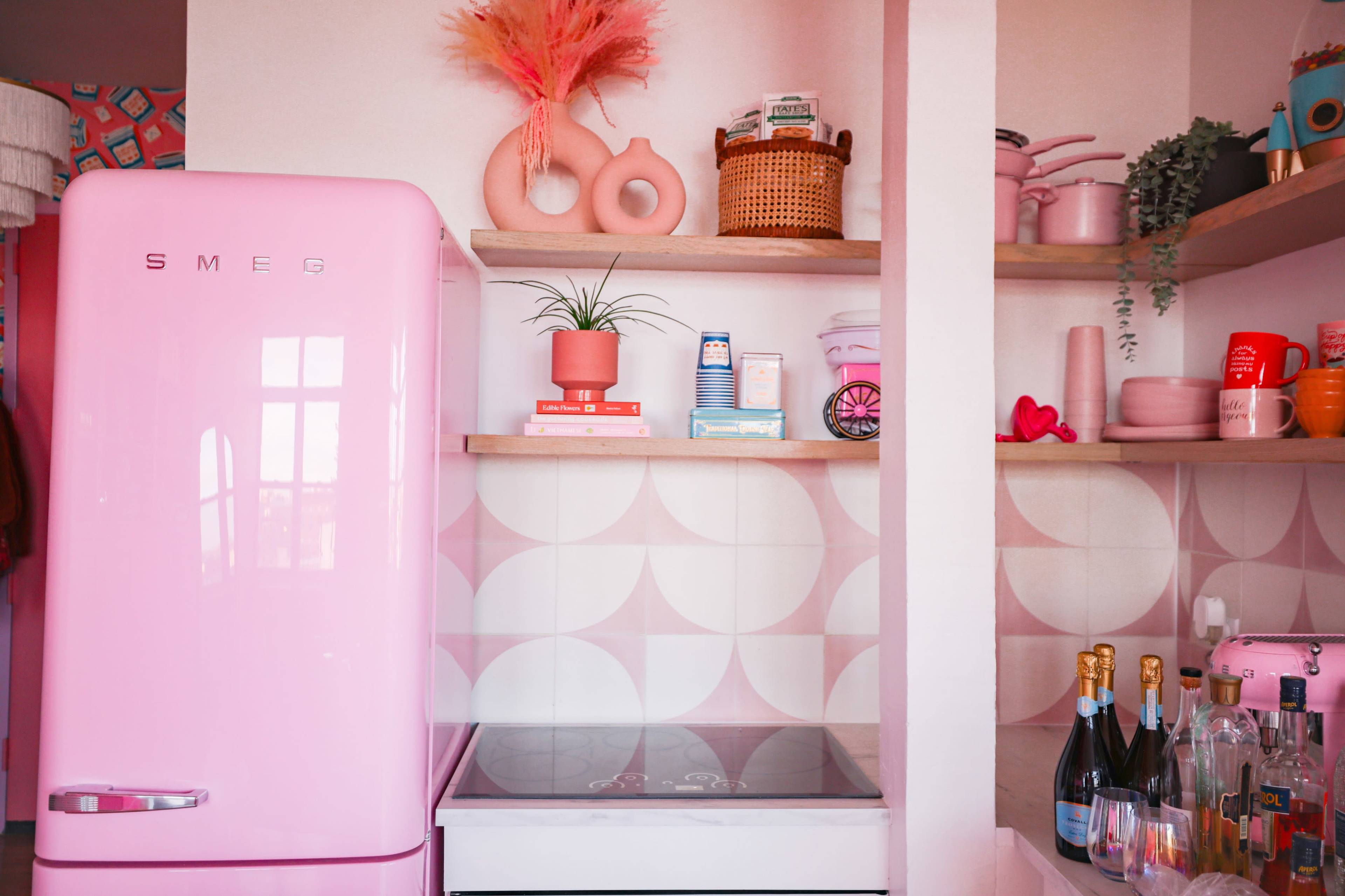 The image features a pink SMEG refrigerator next to shelves displaying various kitchen items, all set against patterned tiled walls.