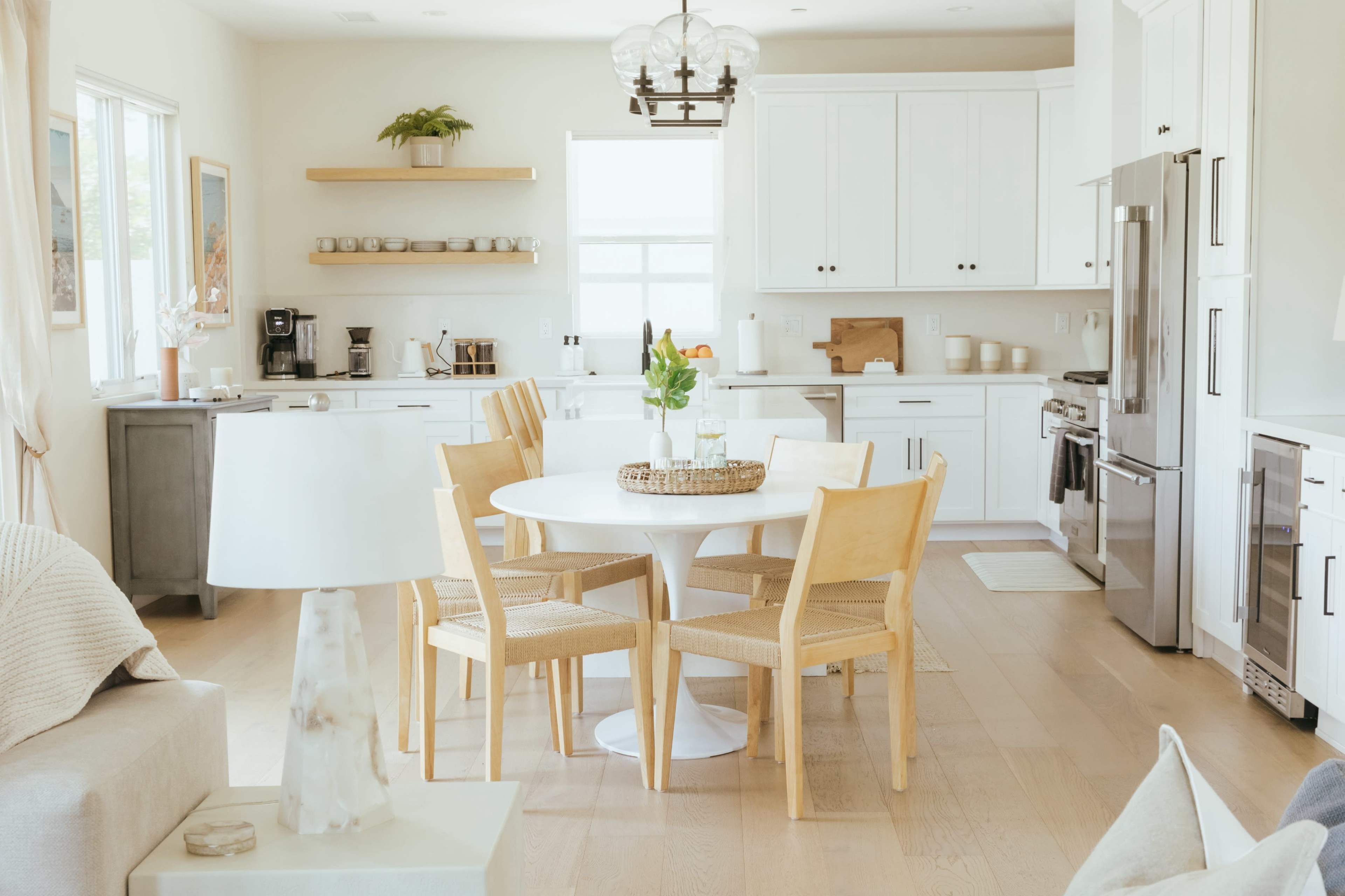 A modern kitchen features a circular white dining table surrounded by four wooden chairs, with cabinets and appliances in the background.