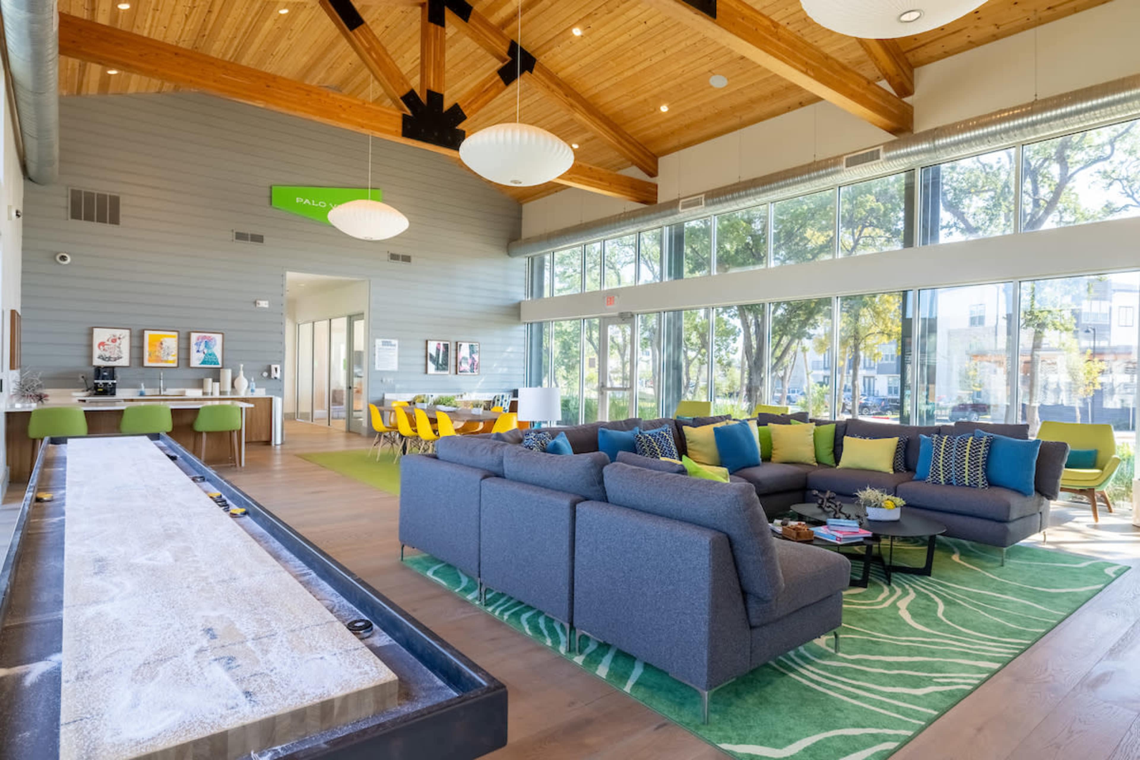 The scene features a spacious community lounge with a wooden ceiling, large windows, a seating area with a sectional sofa, and a long table for social gatherings.