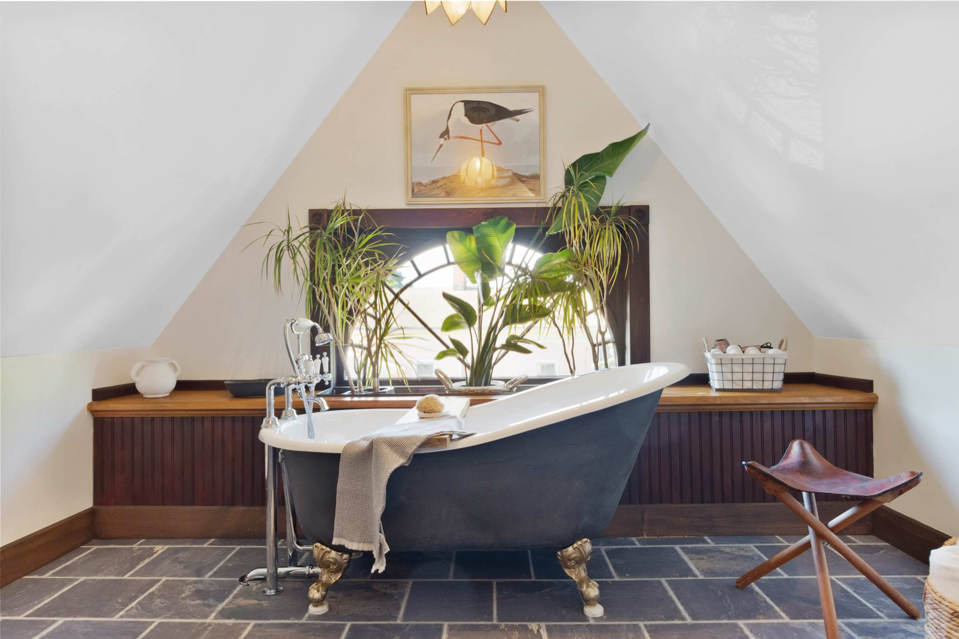 A freestanding bathtub is positioned near a large window surrounded by plants in a well-lit, triangular-shaped bathroom.