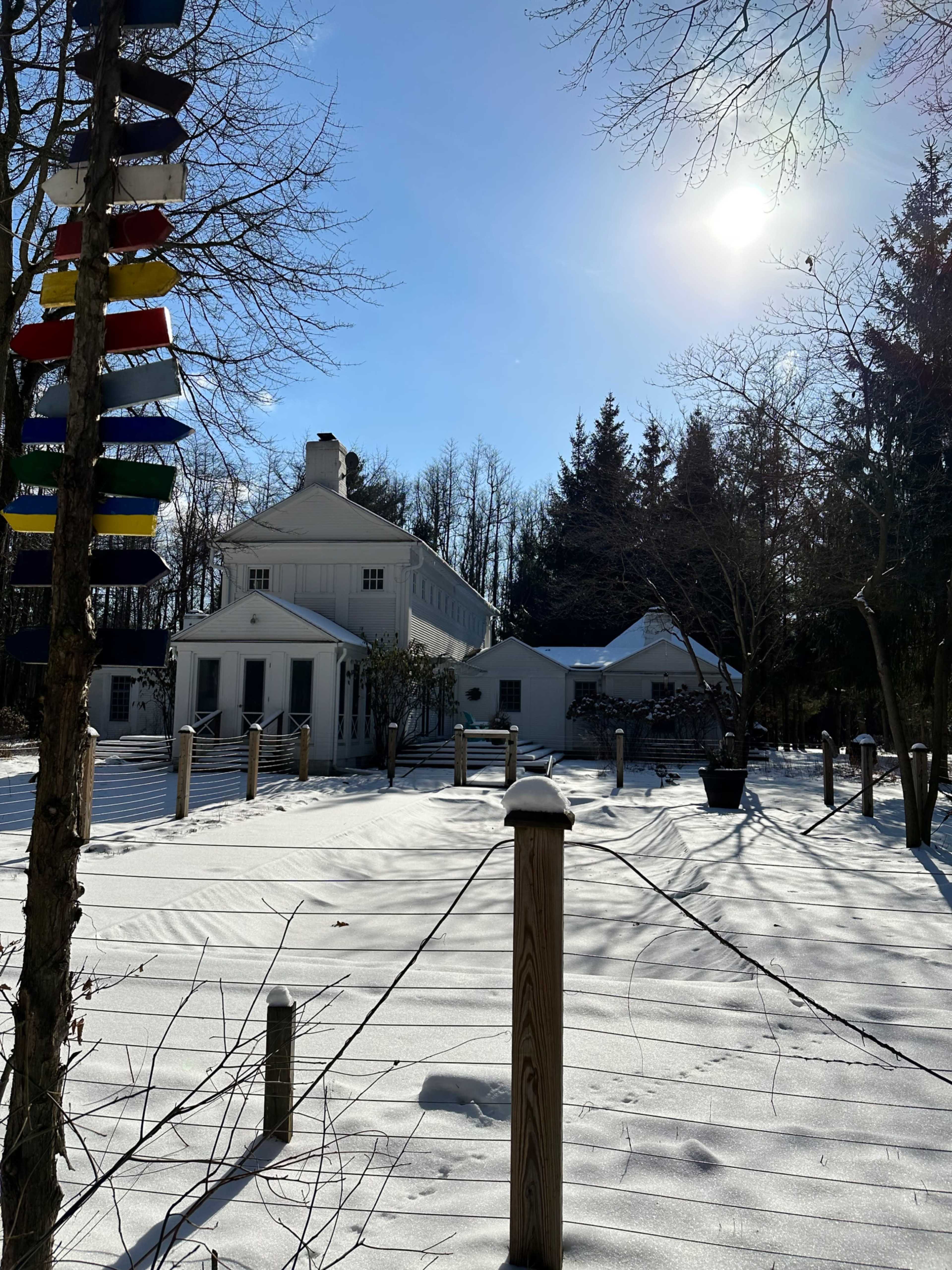 A white house stands in a snowy landscape, with tall trees in the background and colorful flags hanging from a post in the foreground.
