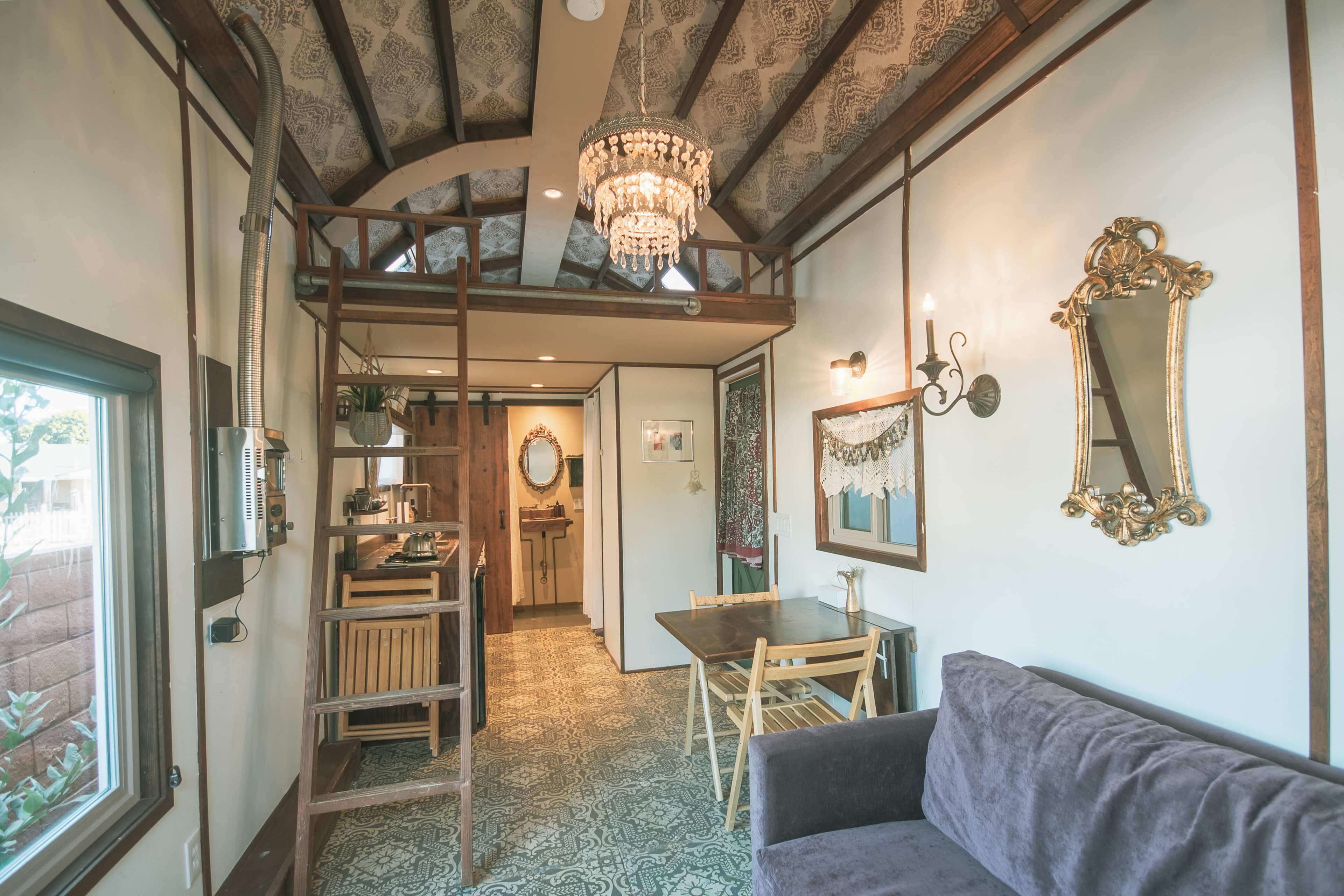 The image shows a cozy interior of a small house featuring a dark sofa, a dining area with wooden furniture, and a staircase leading to a loft, all accented by decorative lighting and ornate details.