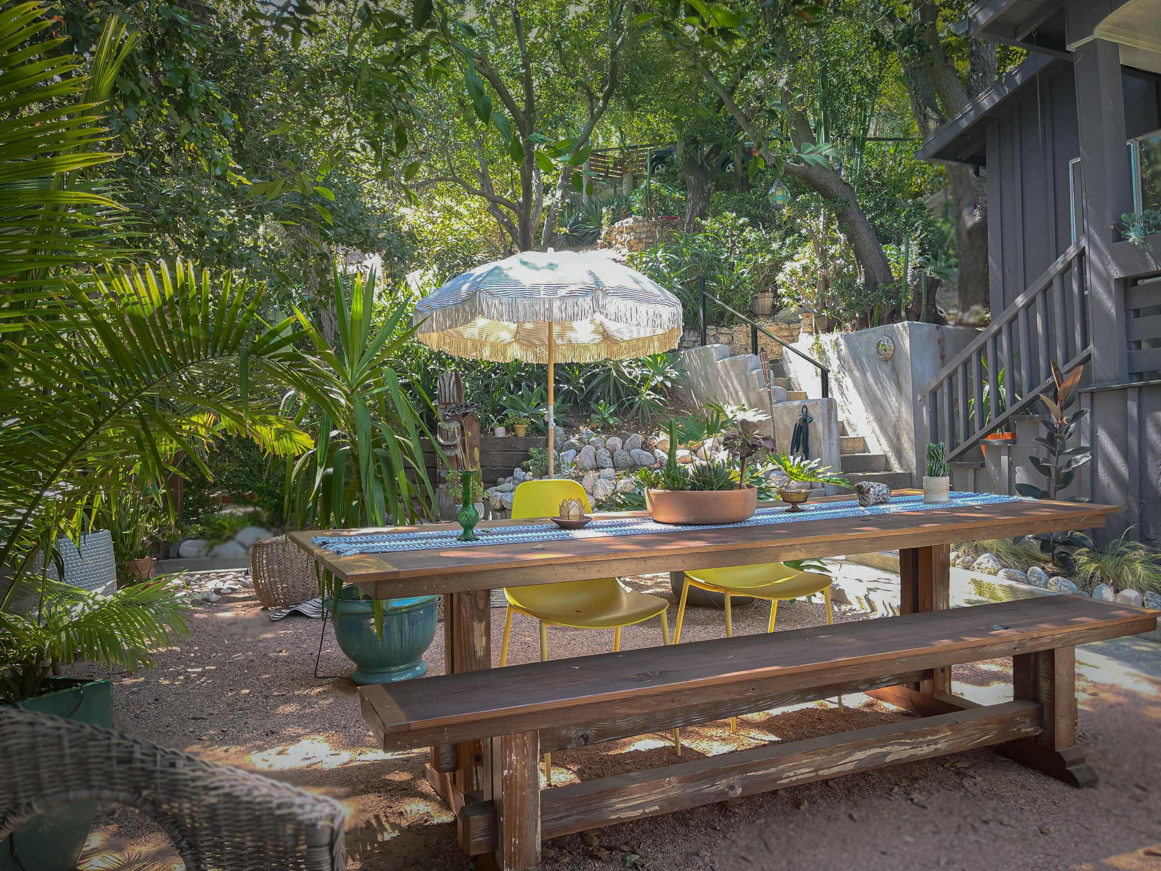 A wooden dining table with yellow chairs sits under a patio umbrella in a lush garden surrounded by greenery and stone pathways.