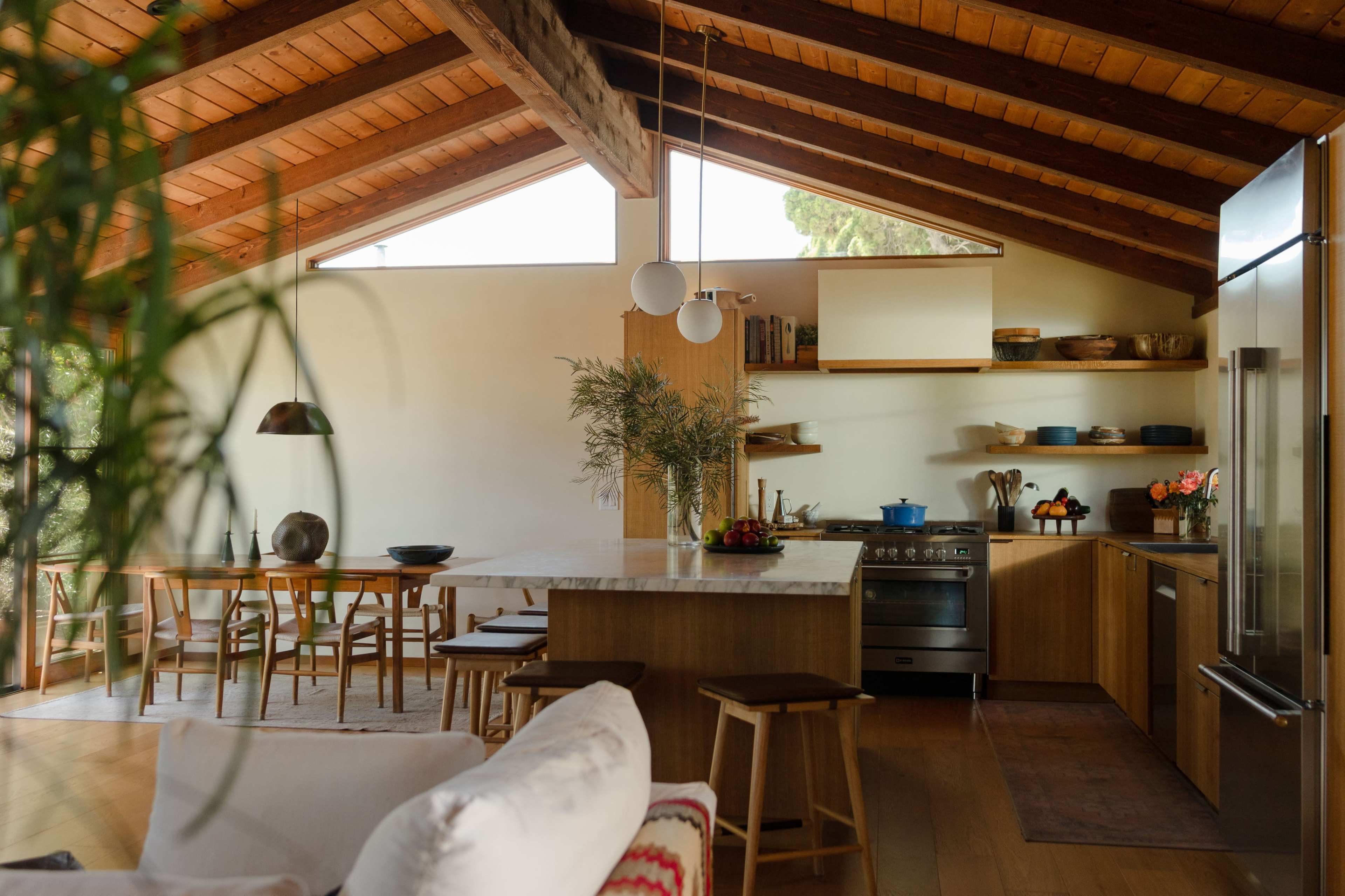 The image shows a modern kitchen with wooden beams, a central island, and an open dining area, featuring a table with chairs and various kitchen accessories.