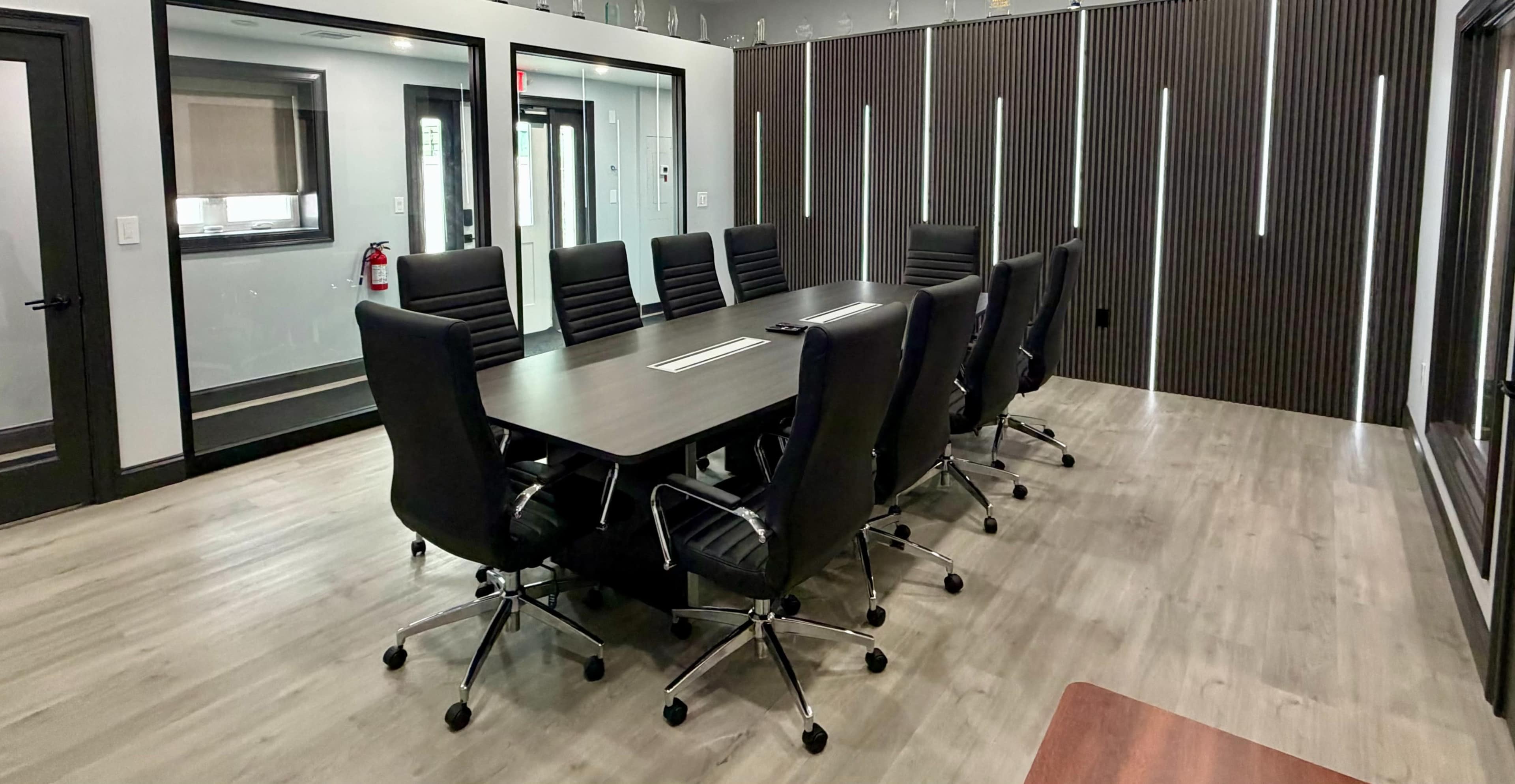 A modern conference room features a large rectangular table surrounded by black ergonomic chairs, with a decorative wall in the background and glass walls on the sides.