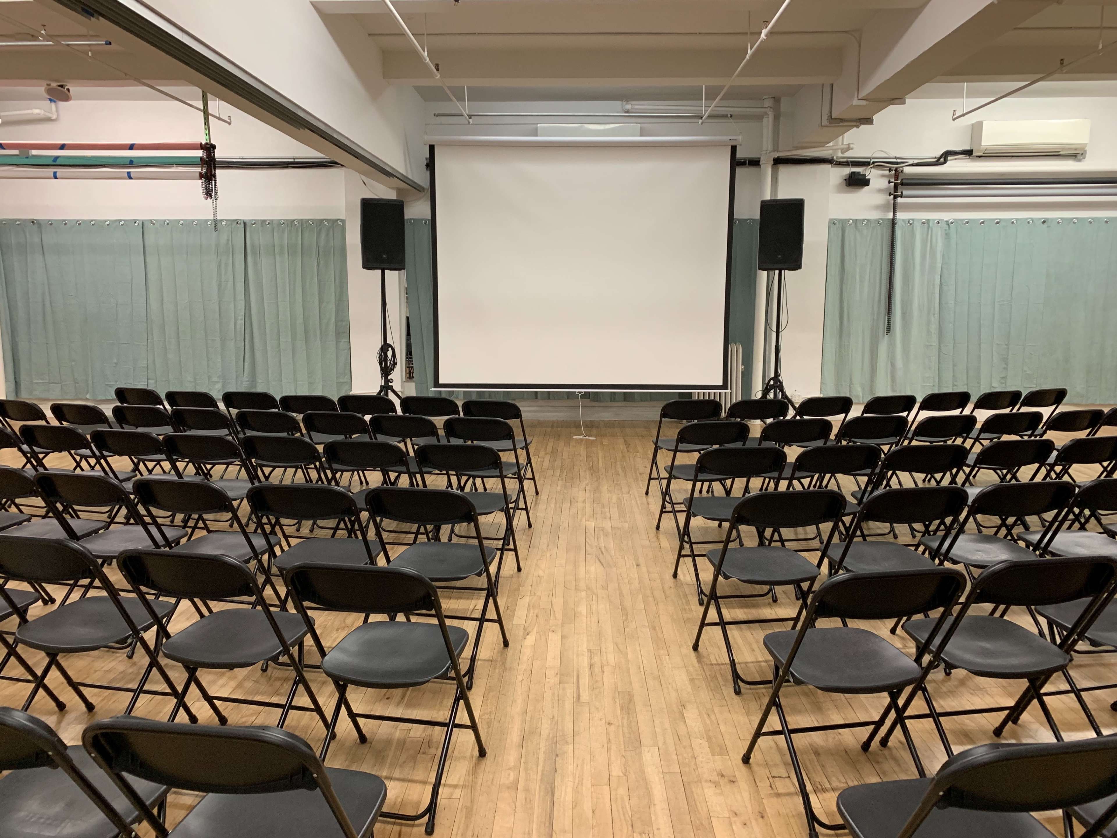 A large, mostly empty room with black folding chairs arranged in rows facing a blank projection screen.