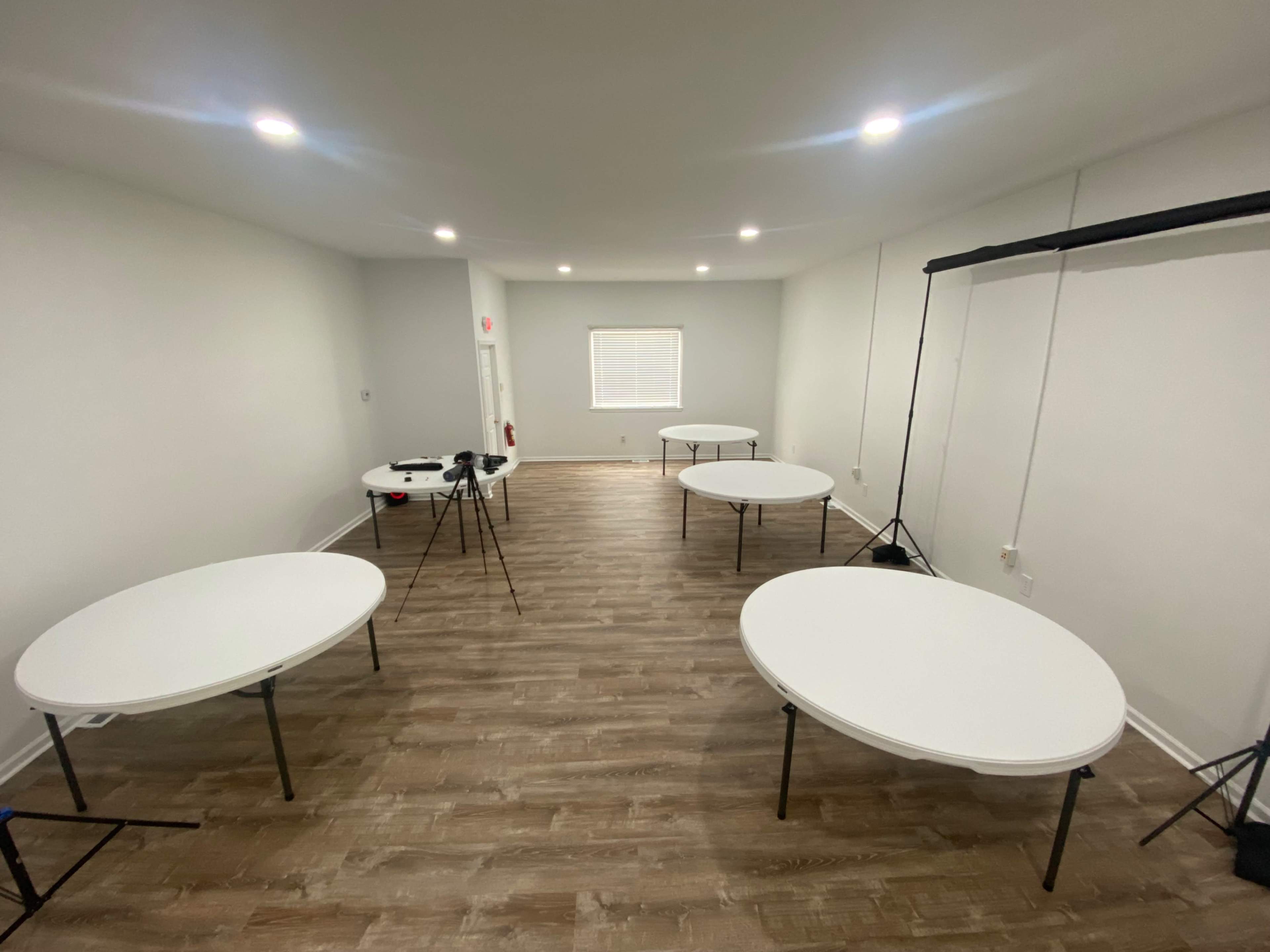 A spacious, empty room with five round white tables arranged around the space and three cameras set up on tripods.