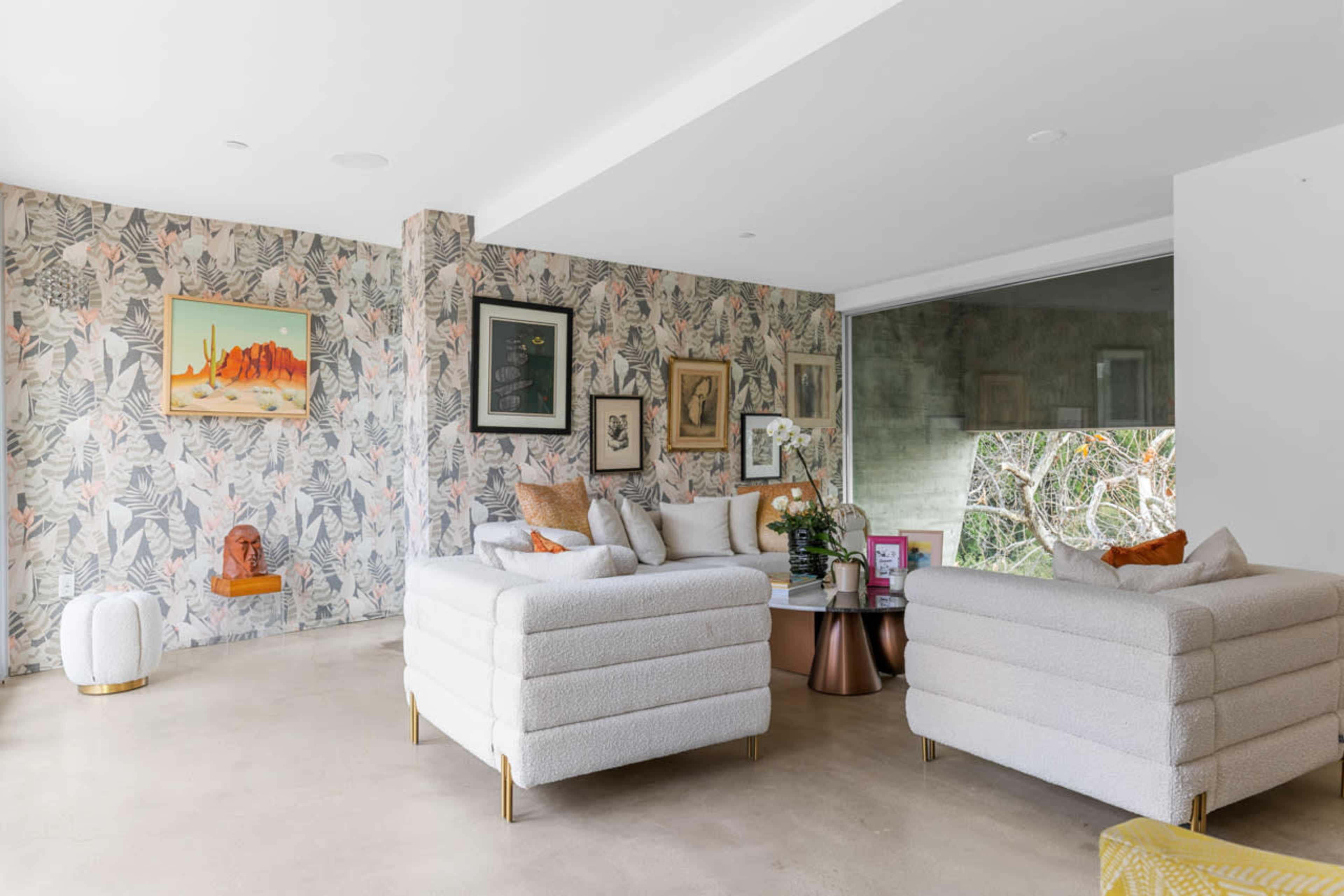 The image shows a modern living room with light-colored sofas, tropical-themed wallpaper, and large windows overlooking a garden.