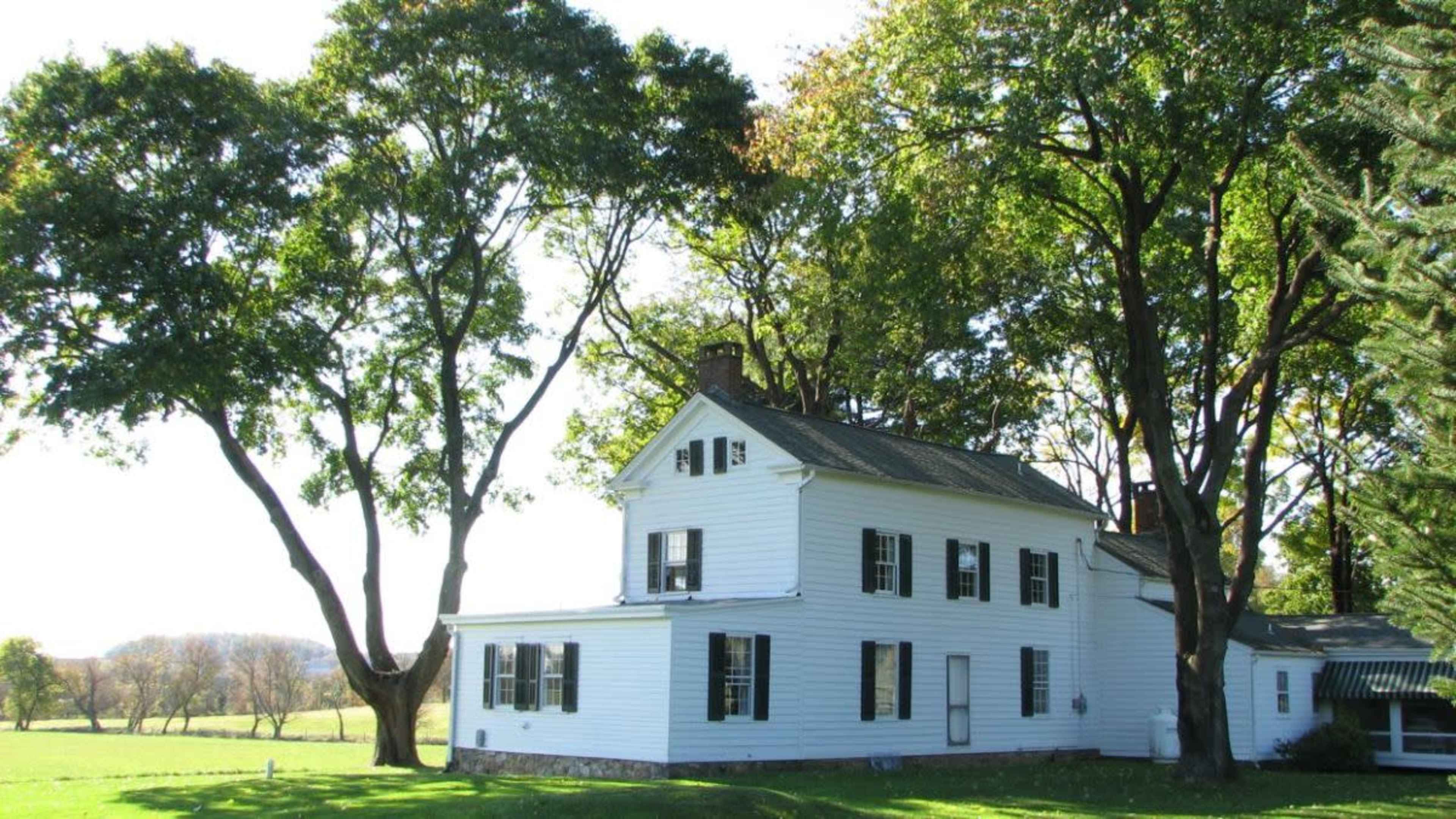 A white house with black shutters sits on a grassy area surrounded by large trees.