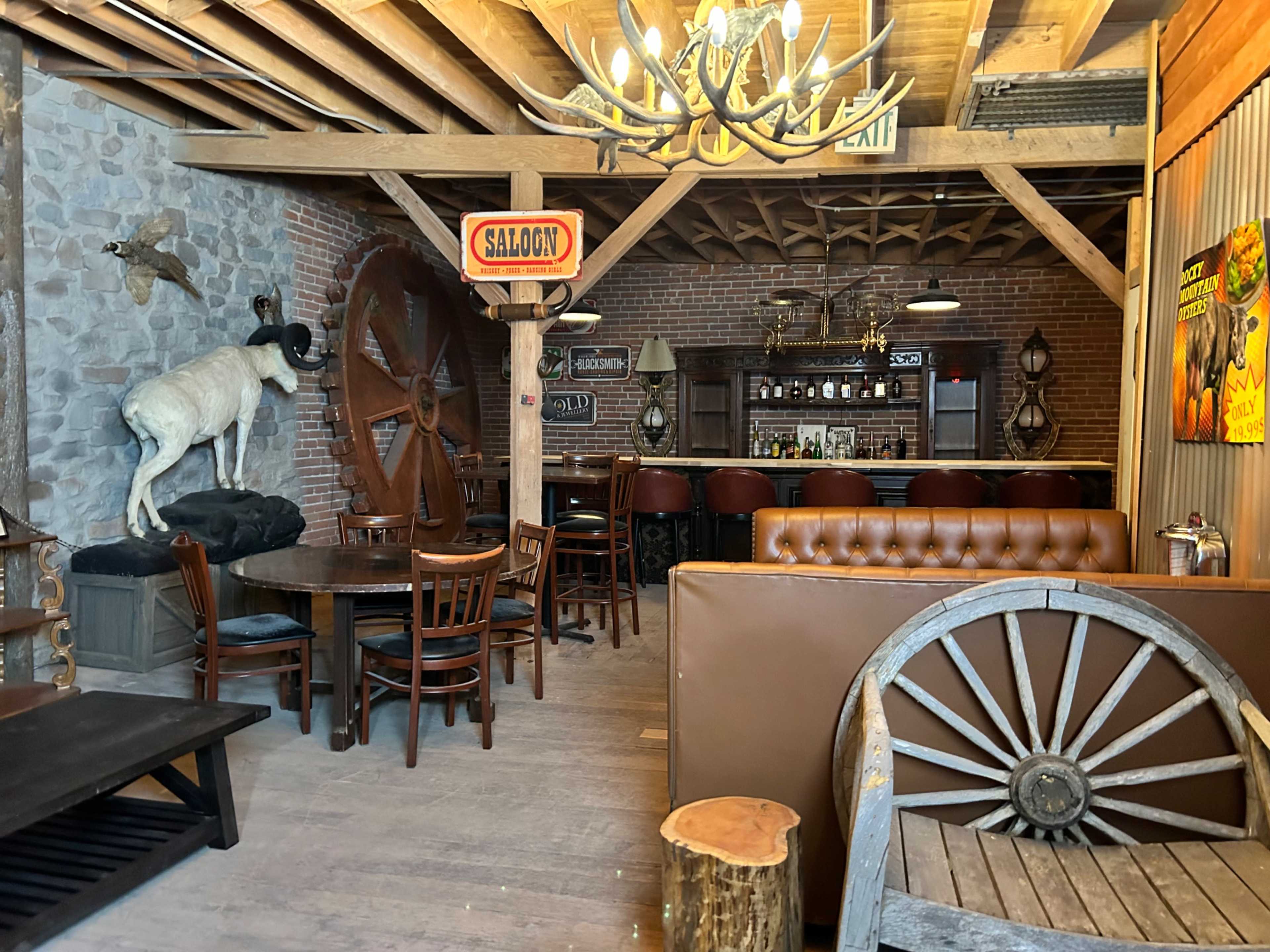 The interior of a rustic saloon features wooden beams, a bar lined with stools, a dining area with wooden tables, and decor including a mounted animal and an old wagon wheel.