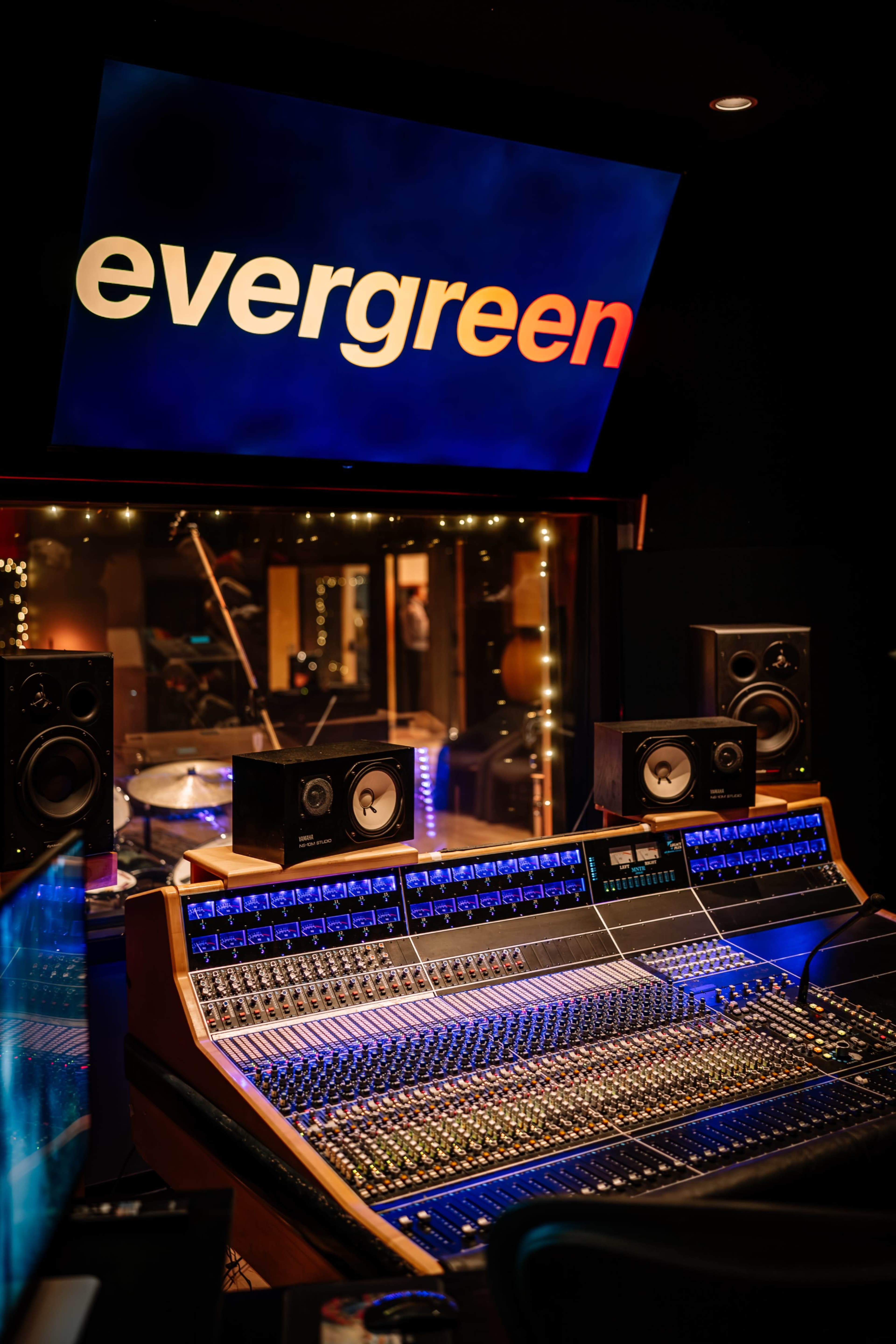 The image shows a music recording studio with a large mixing console in the foreground and a lit sign displaying the word "evergreen" in the background.